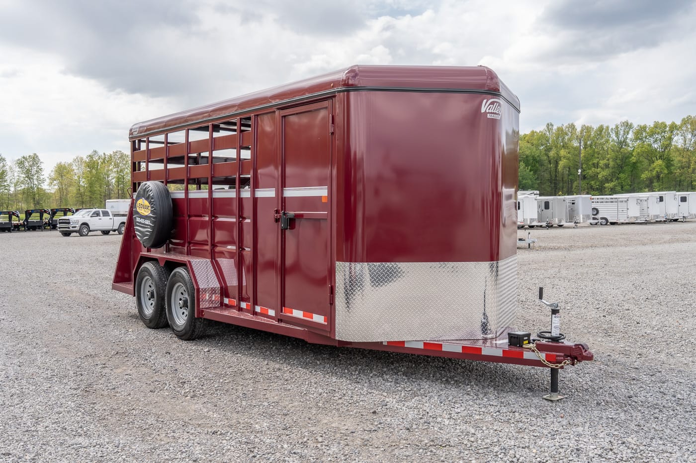 Maroon two-axle livestock trailer with spare tire mounted on the side, parked on a gravel lot.