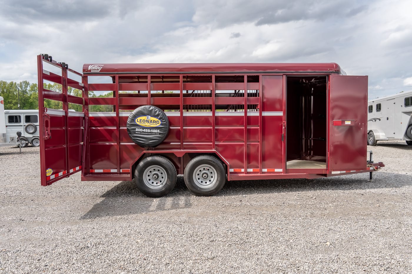 Red livestock trailer with dual axles and open side access doors on a gravel lot, rear door ajar.