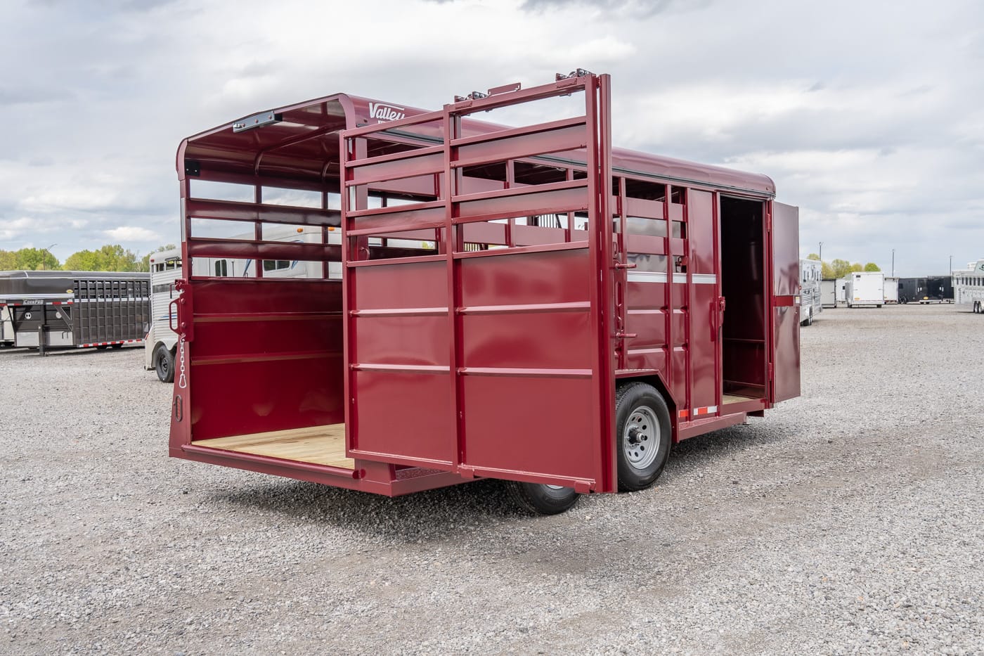 Red metal livestock trailer with side doors open, parked on a gravel lot, showing a wooden floor interior and rear ramp area on a cloudy day