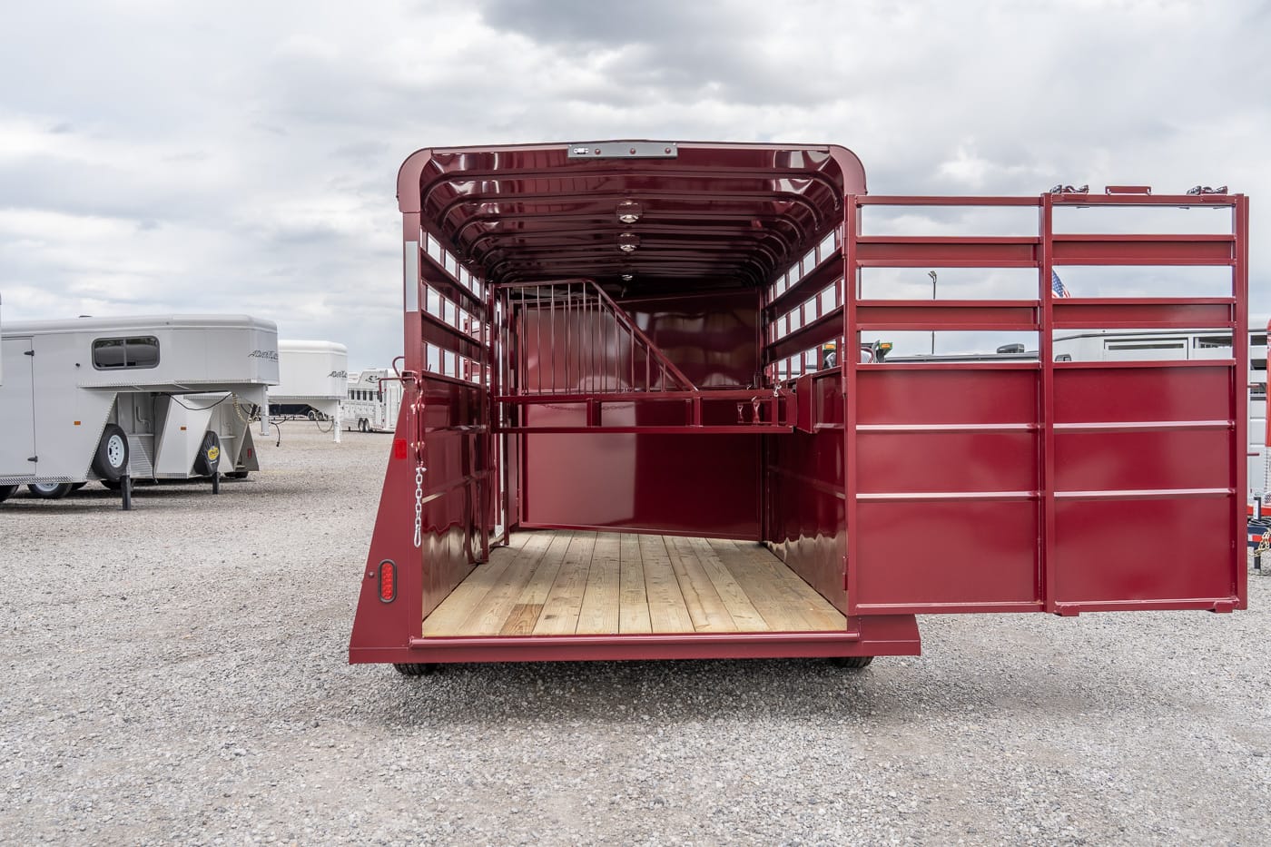 Open-back maroon livestock trailer with wooden floor and center divider in a gravel lot (other trailers in background).