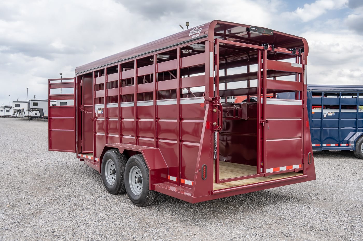 Red two-axle livestock trailer with its rear gates open, parked on a gravel lot, showing a wooden floor interior.