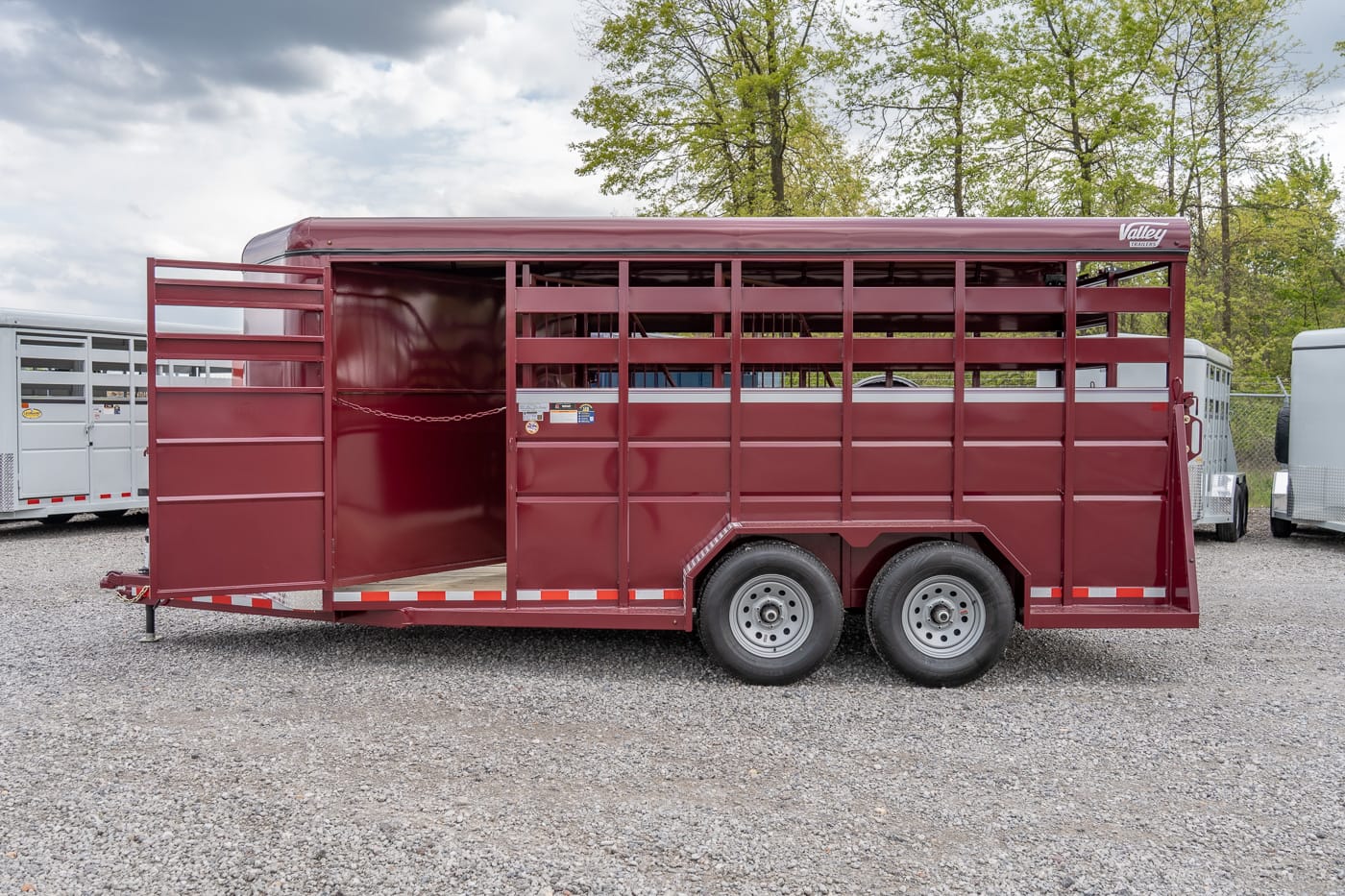 Maroon two-axle livestock trailer with open side rails on a gravel lot.