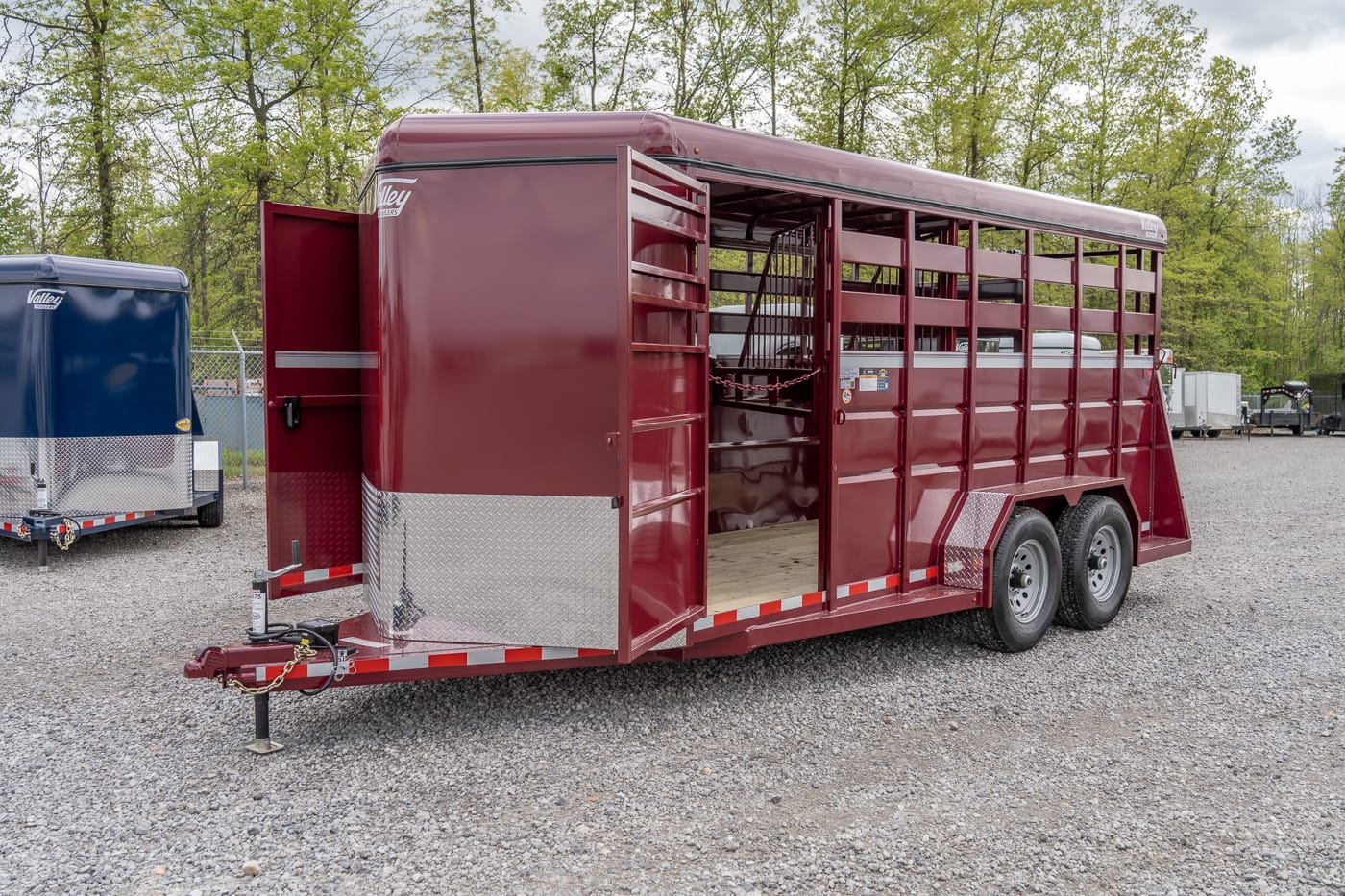 Maroon livestock trailer with open side door, two axles, and diamond-plate trim parked on a gravel lot under trees.