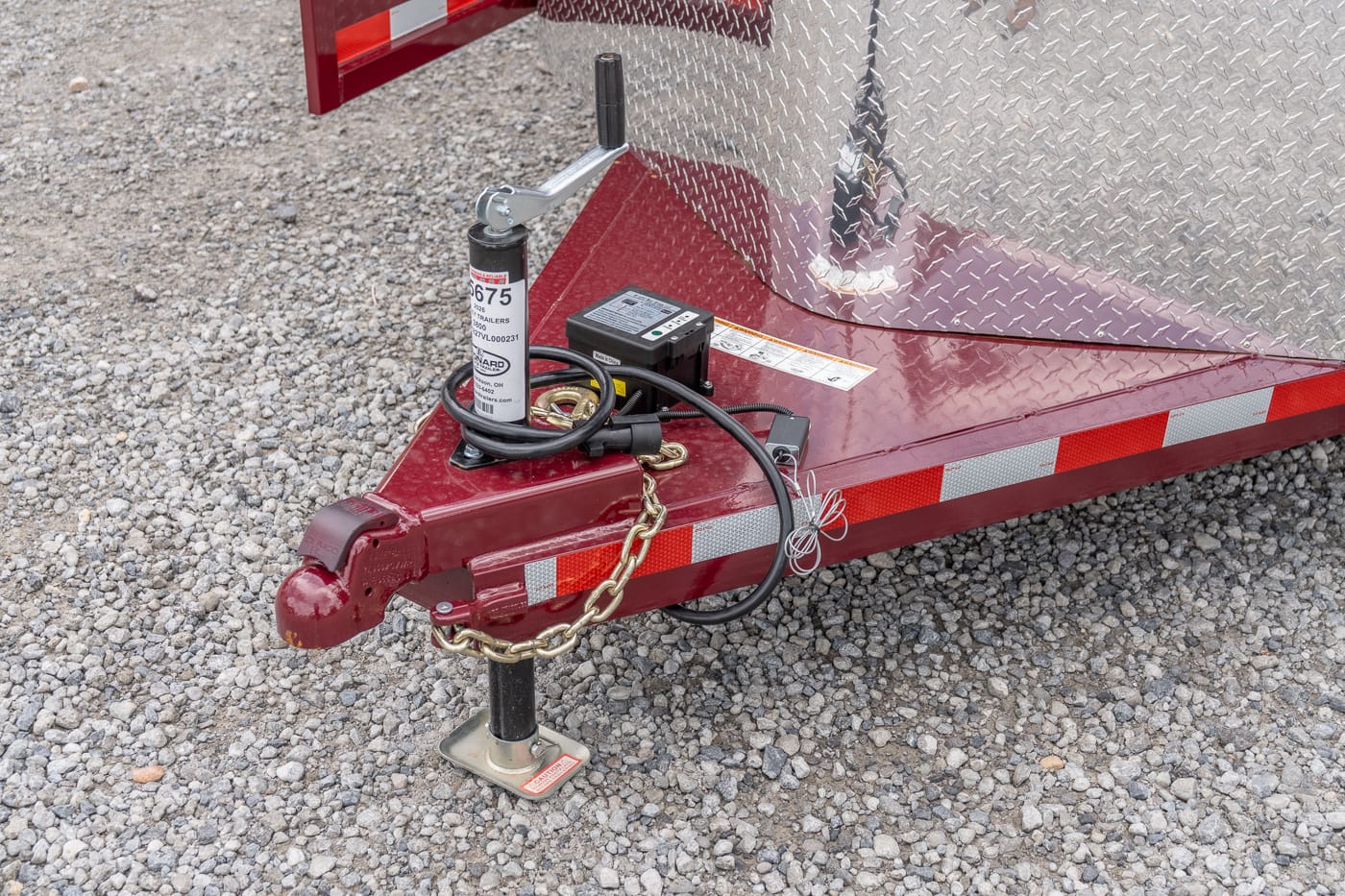 Close-up of a red trailer tongue on gravel, showing a manual jack, safety chain, wiring, and a diamond-plate fender area in the background