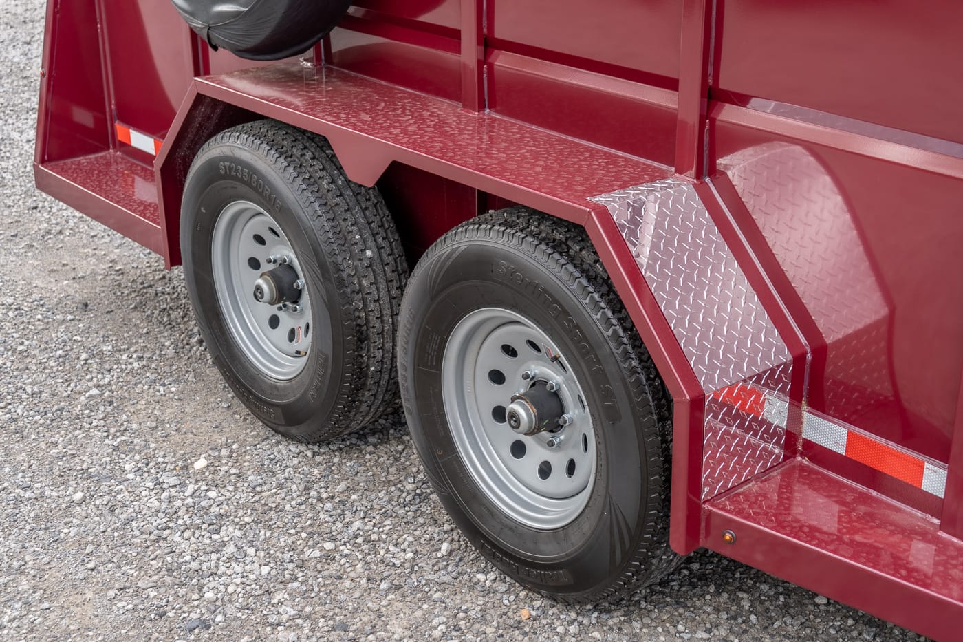 Close-up of two rear wheels on a red utility trailer with diamond-plate fenders on gravel ground.