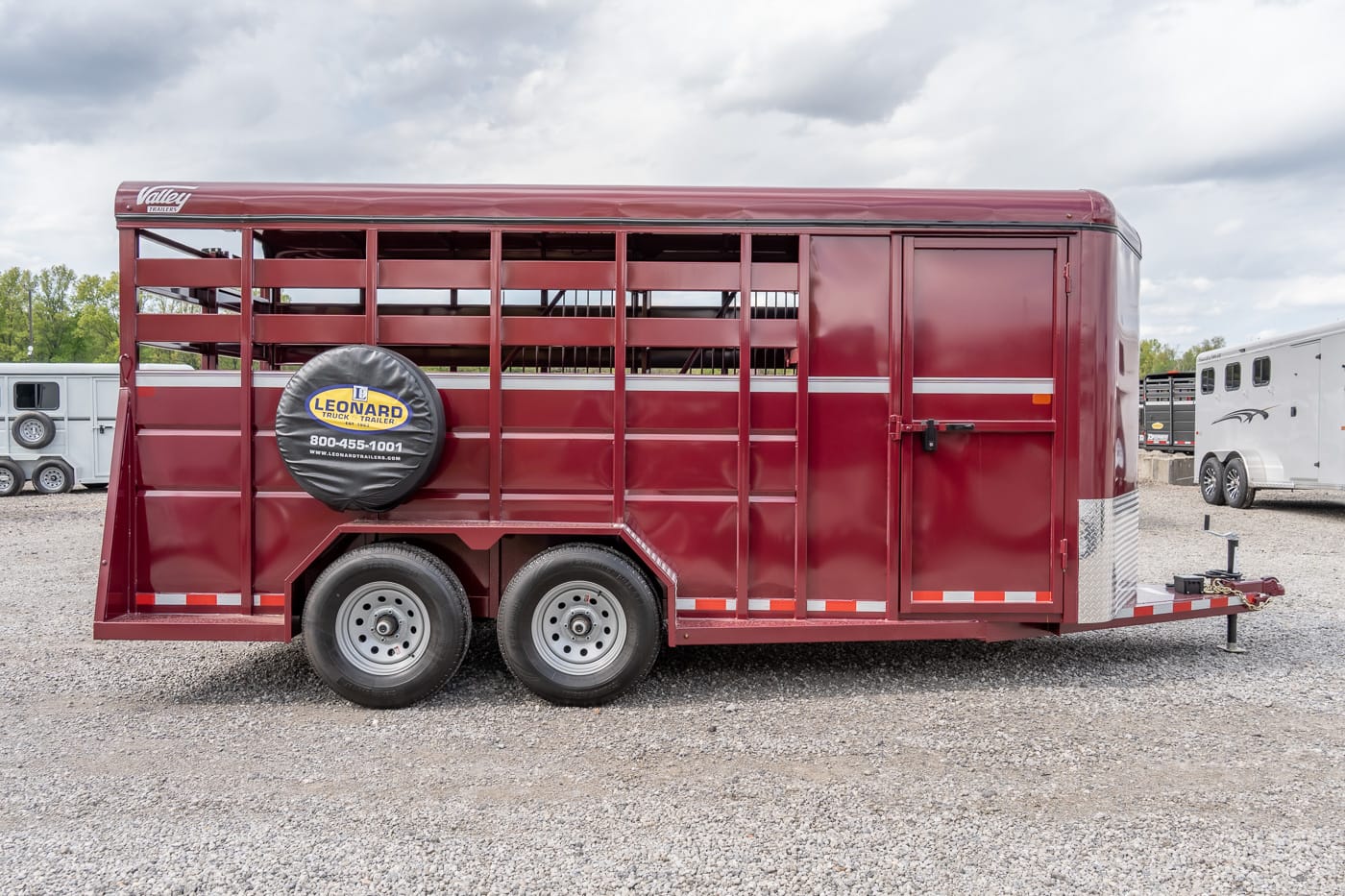 Red metal horse trailer with two axles and a spare tire mounted on the side, parked on a gravel lot with other trailers in the background.