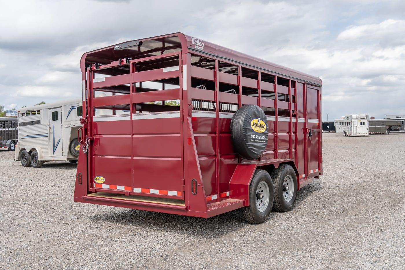 Red maroon two‑axle livestock trailer with a side-mounted spare tire on a gravel lot, other trailers visible in the background.