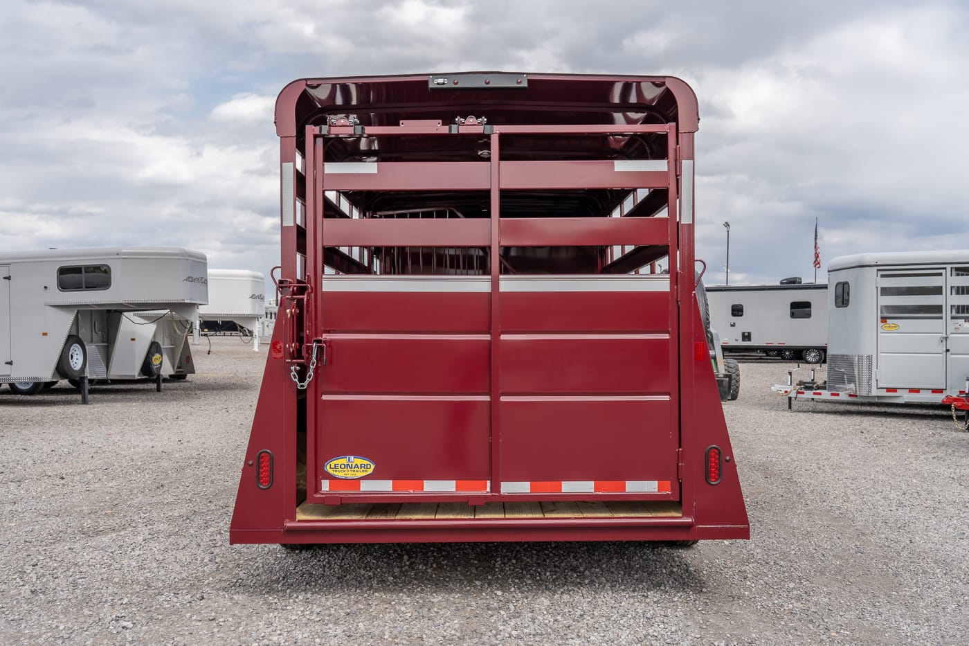 Rear view of a red livestock trailer with open slatted stalls on a gravel lot, ready for loading animals.