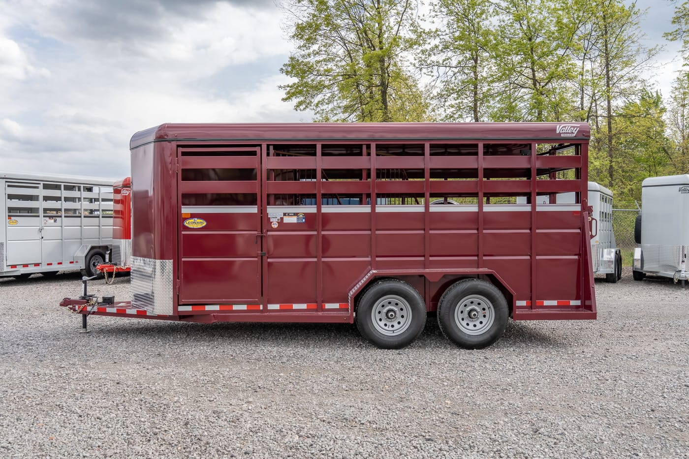 Maroon, two‑axle livestock trailer with vented side slats on a gravel lot, with other trailers nearby and trees in the background.