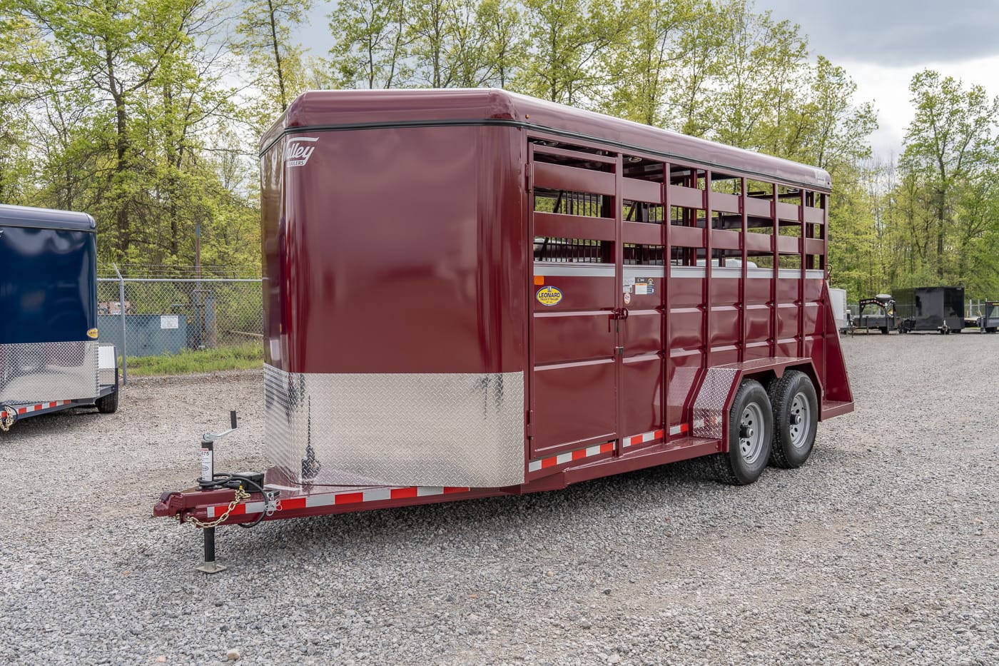 Maroon two‑axle horse trailer with diamond-plate front, parked on a gravel lot amid trees.