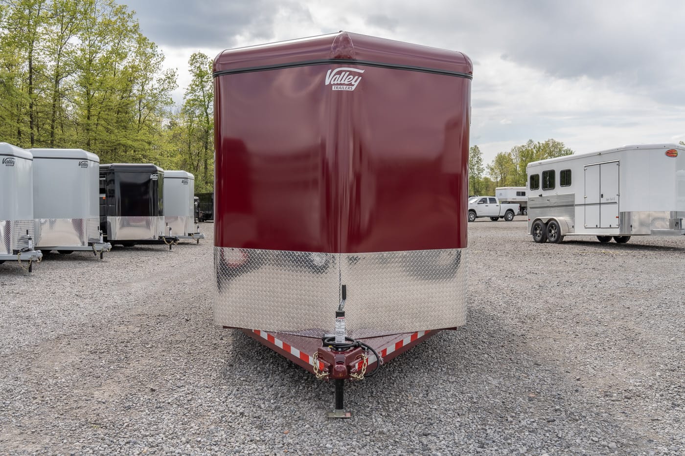 Maroon enclosed trailer with diamond-plate front, hitch attached, parked among other cargo trailers on a gravel lot.