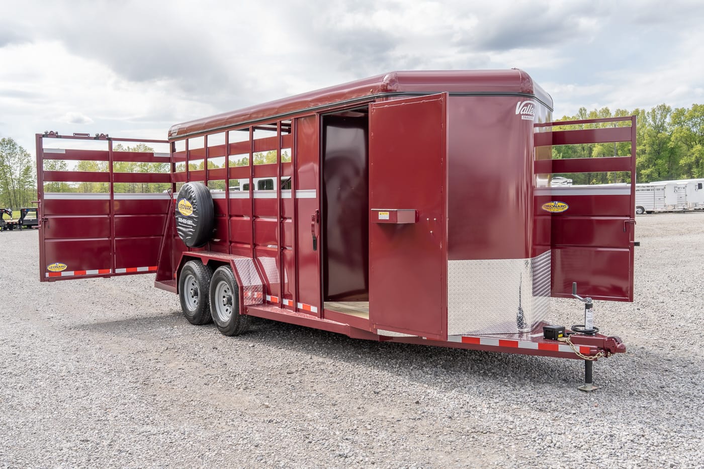 Red livestock trailer with open side access doors, spare tire mounted on the frame, and tandem wheels in a gravel lot.