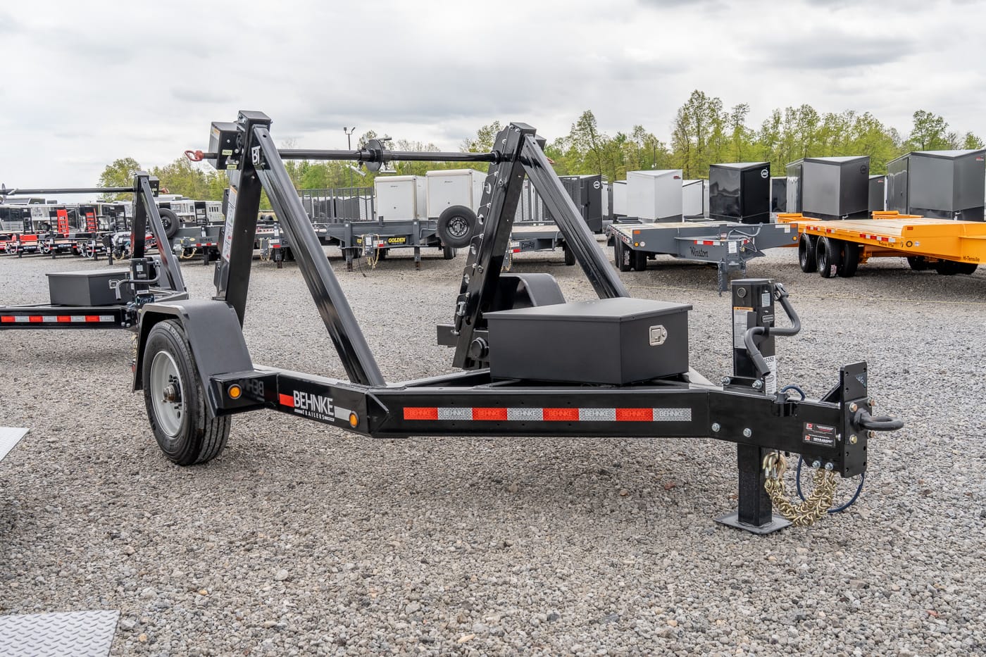 Black dual-axle gooseneck trailer with folding arms and a top toolbox, parked on gravel in a trailer yard.