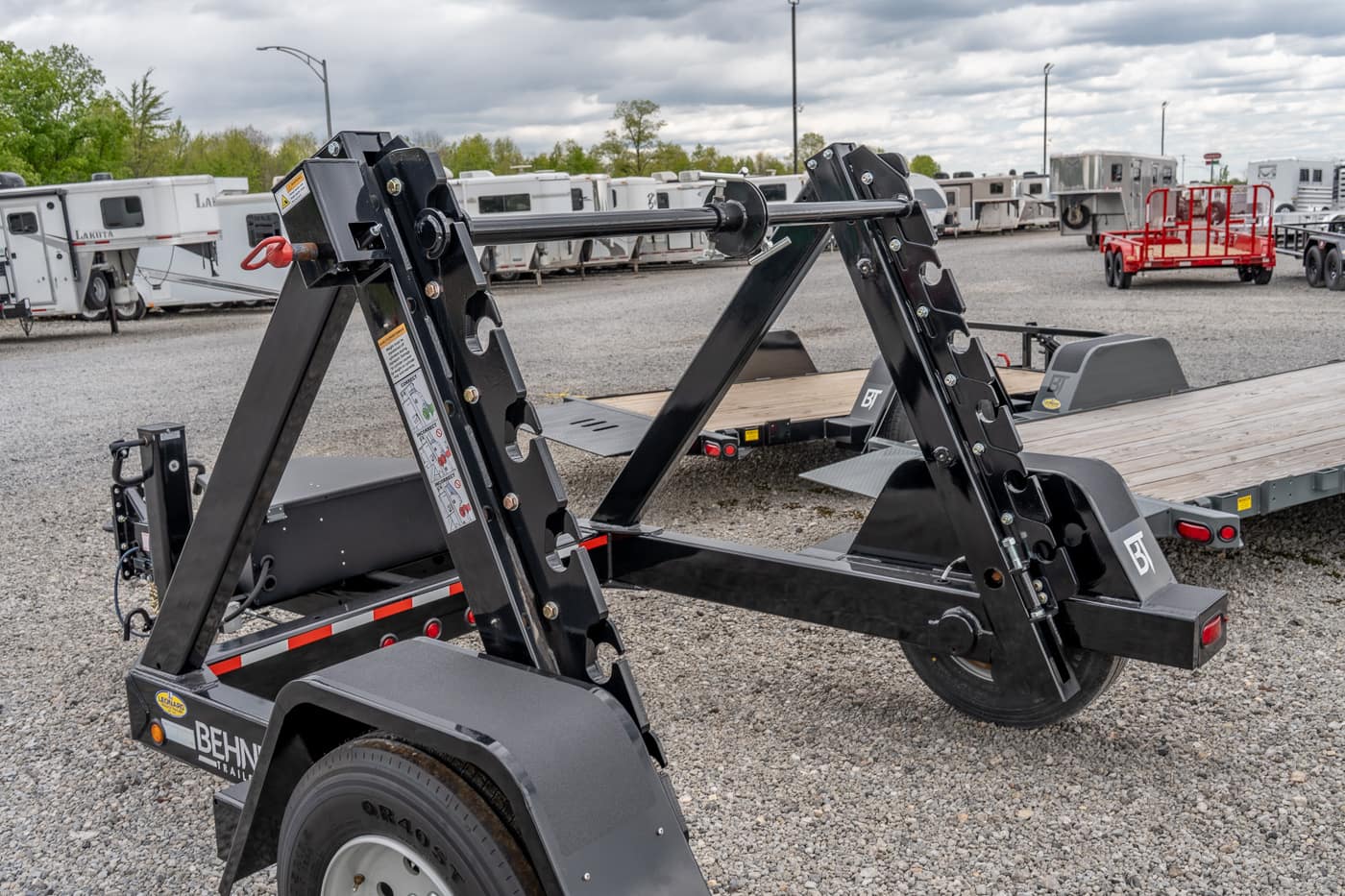 Heavy-duty black dual-axle trailer frame with adjustable bunk supports on a gravel lot, surrounded by other trailers at a dealership.