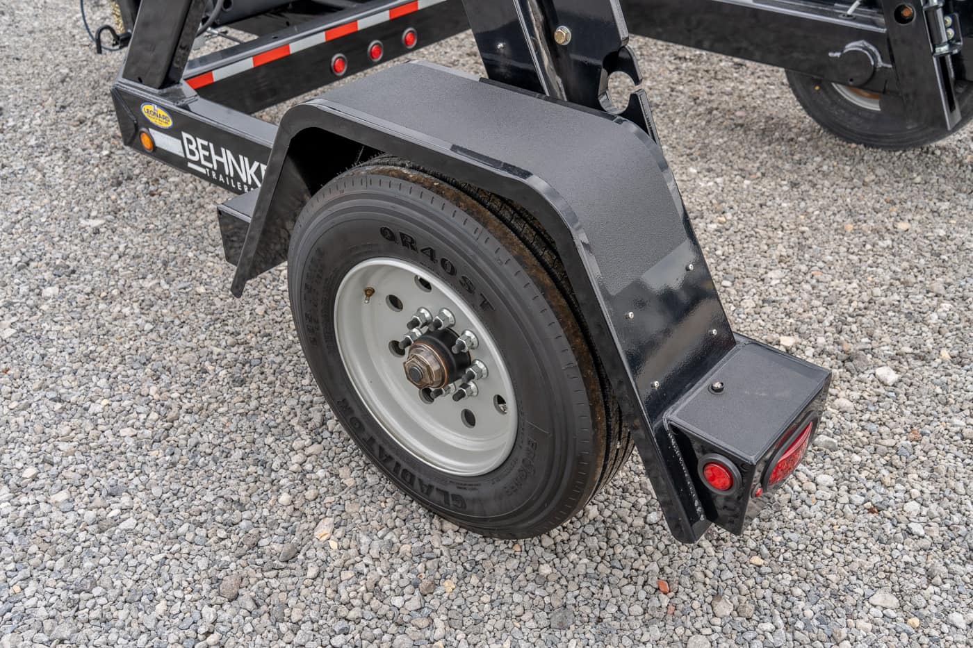 Trailer wheel and black fender on a gravel surface, showing axle hub and stud bolts with a red taillight nearby on the trailer.
