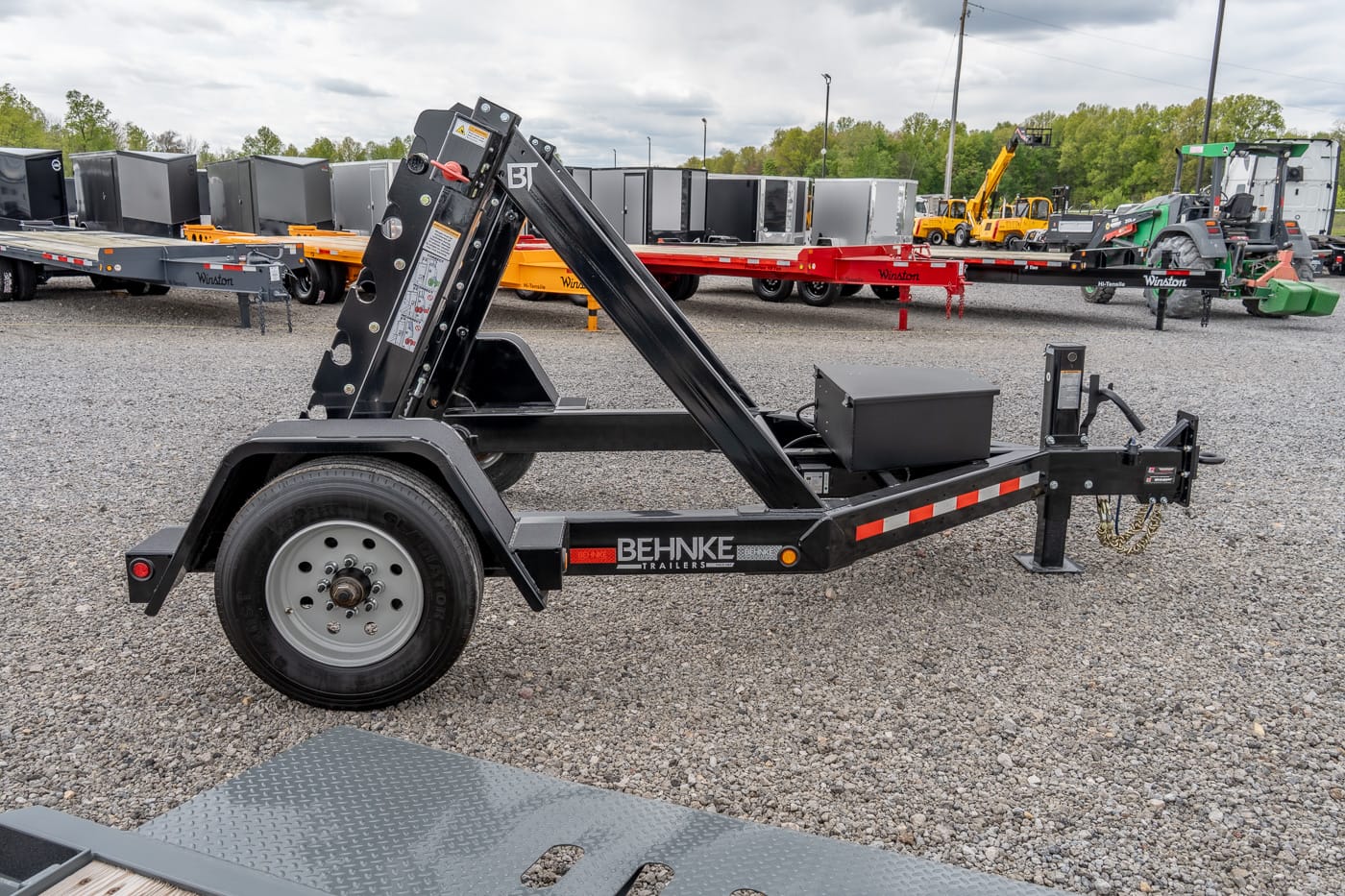 Black Behnke trailer with a hydraulic lifting arm in a gravel lot among other trailers and equipment, ready for use as a tilt-bed loader.