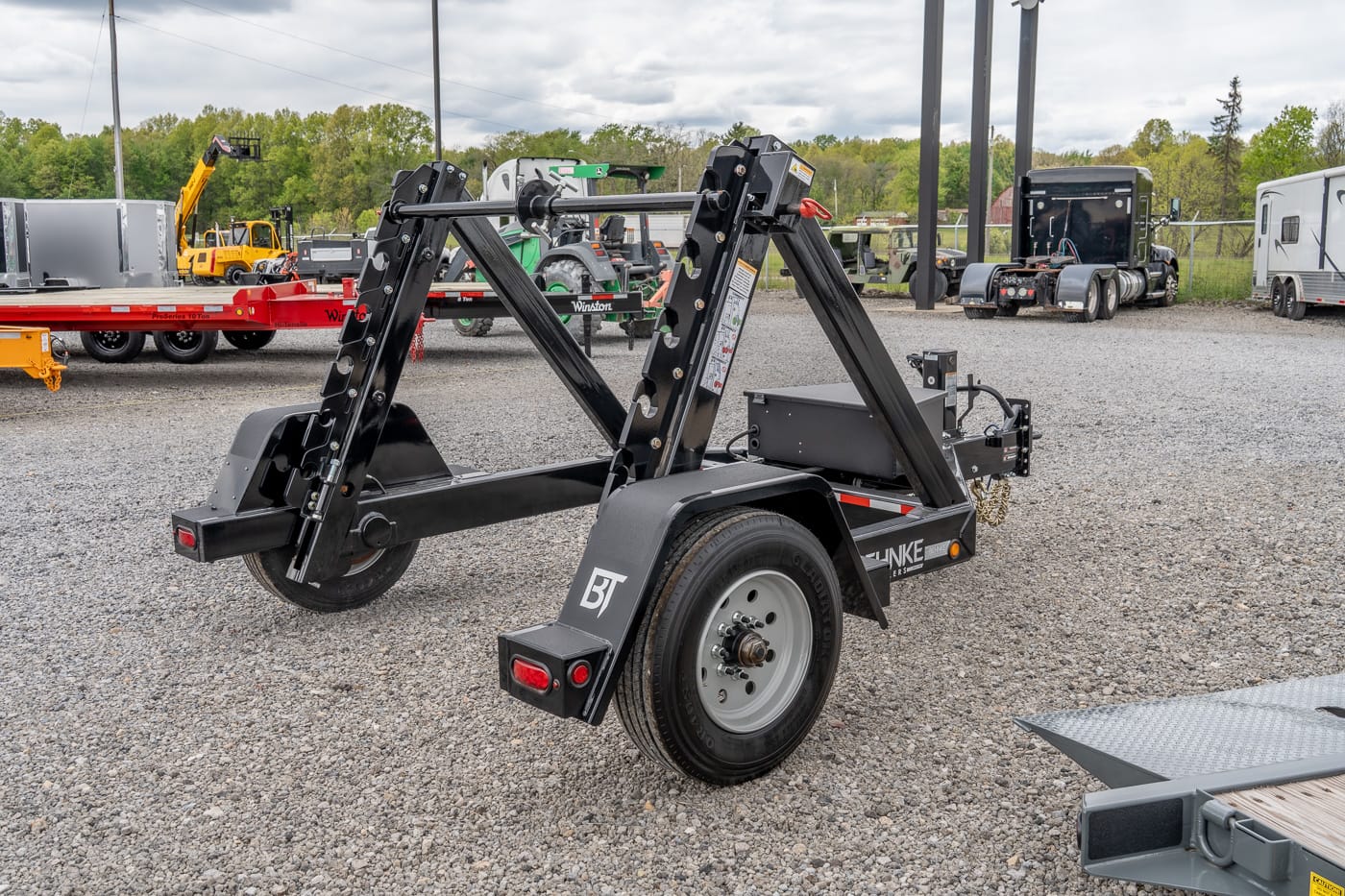 Black hydraulic tilt trailer with dual lifting arms and a mounted spare wheel in a gravel lot, ready for transporting heavy equipment.