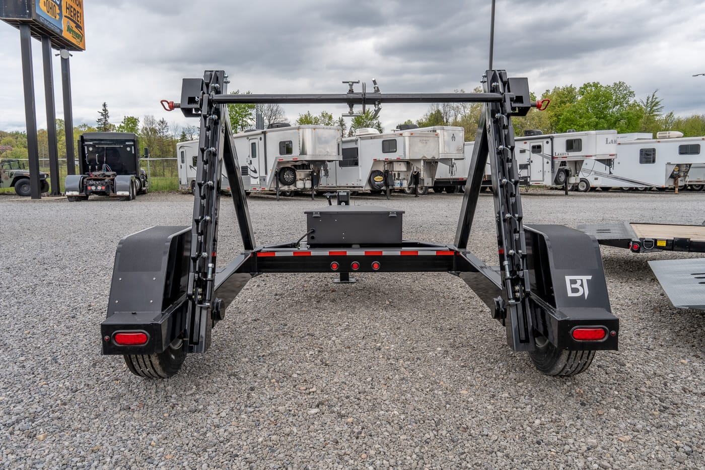 Rear view of a black BT-brand heavy-duty trailer frame with dual angled arms, in a gravel lot with RVs in the background.