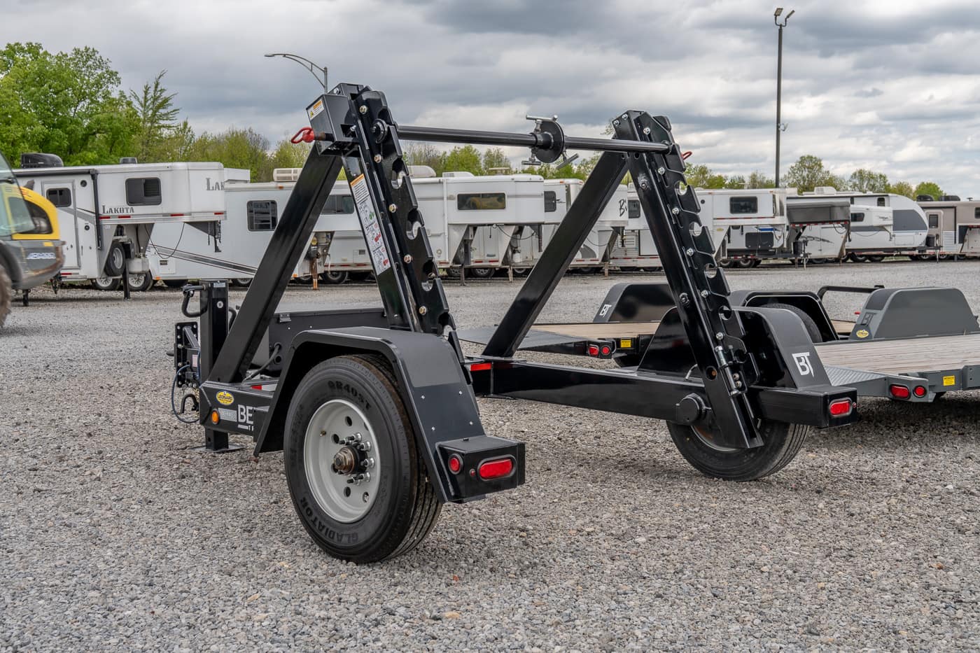 Black heavy-duty trailer with folding dual arms and single wheel on a gravel lot, parked among RVs and campers in the background, used for vehicle loading.