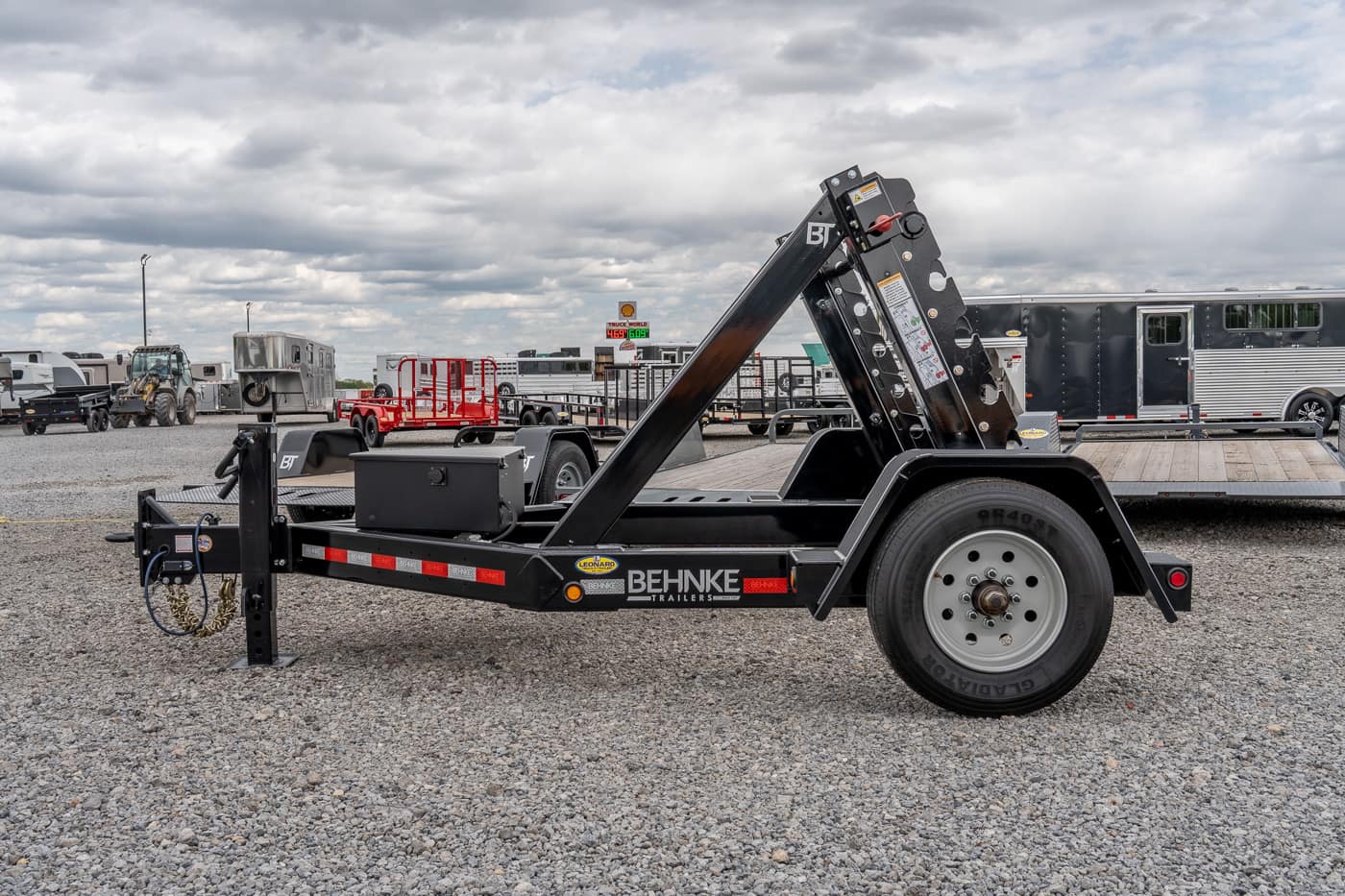 Black tilt-bed trailer with hydraulic arm and Behnke Trailers logo, parked on a gravel lot with other trailers nearby.