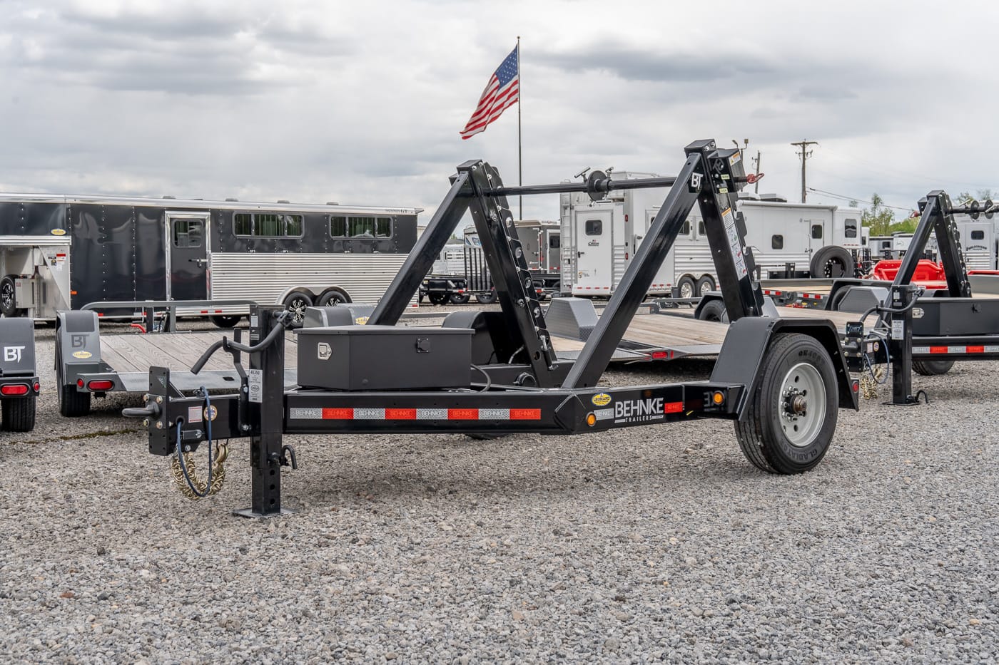 Black Behnke trailer with folding dual-arm ramps on a gravel lot, American flag overhead, other trailers and horse trailers in the background.