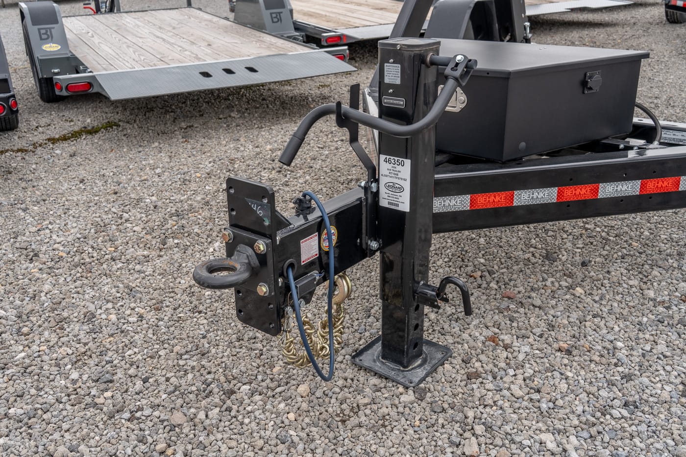 Close-up of a trailer tongue with a manual jack, safety chains, and a black storage box on a gravel lot