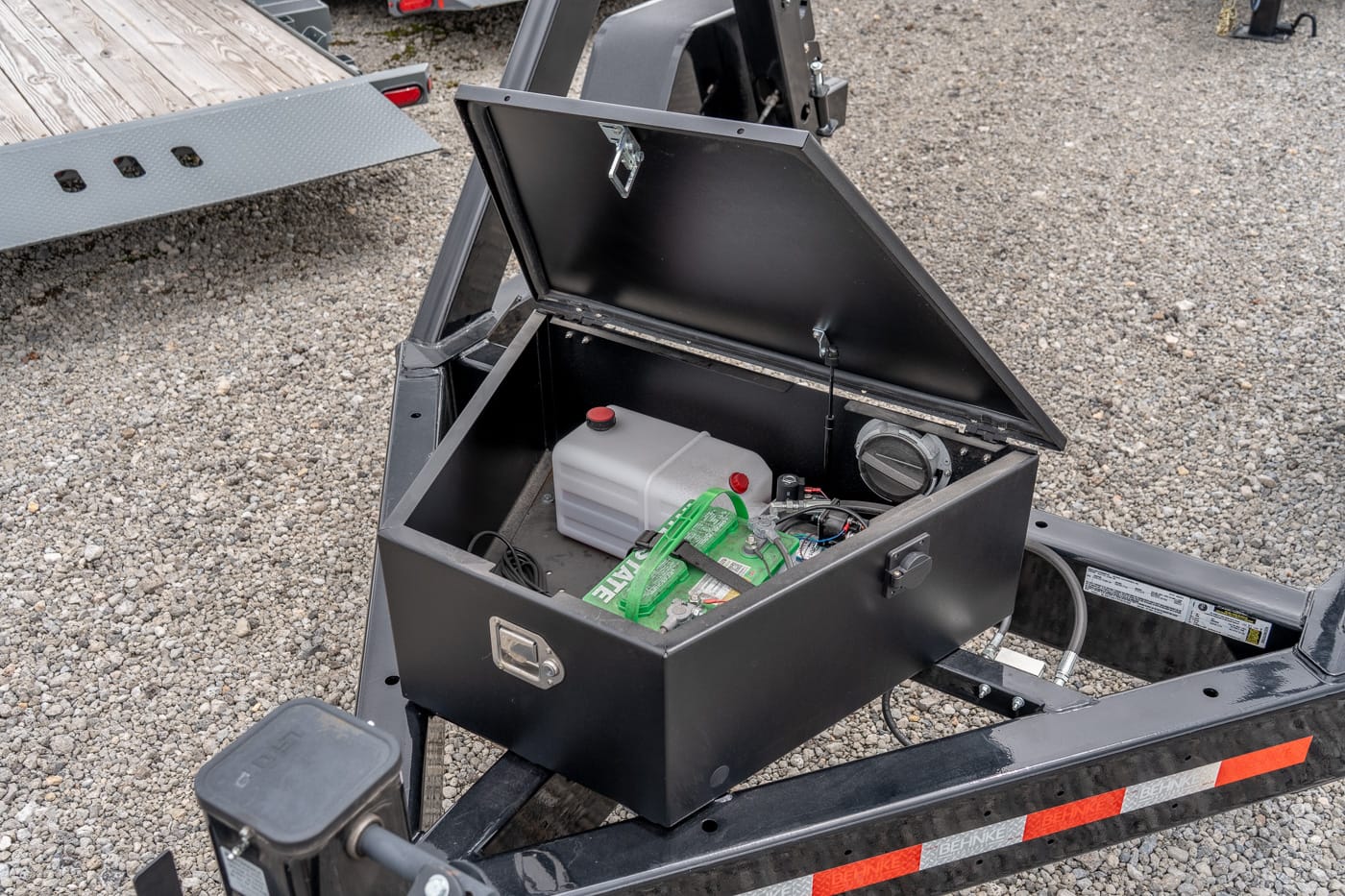 Open black metal toolbox on a trailer frame, containing a white plastic reservoir, a car battery, and assorted cables.