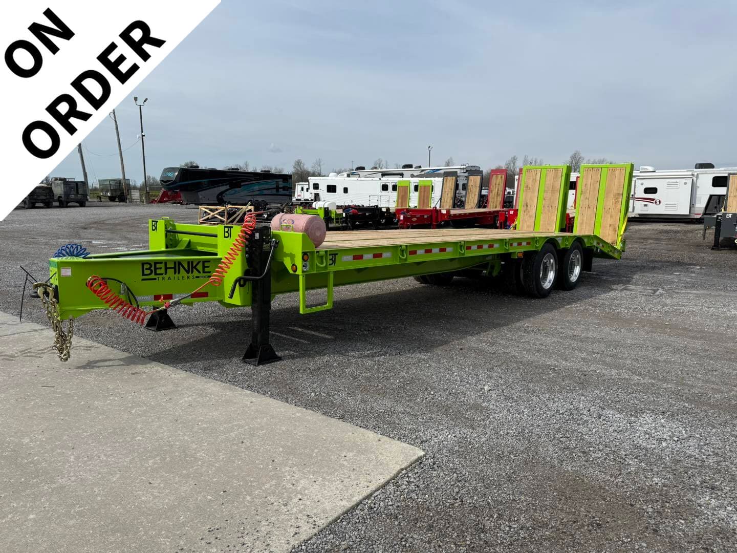 Bright green Behnke flatbed trailer with wooden deck and loading ramps on a gravel lot, RVs in background, 'ON ORDER' banner diagonally across corner.