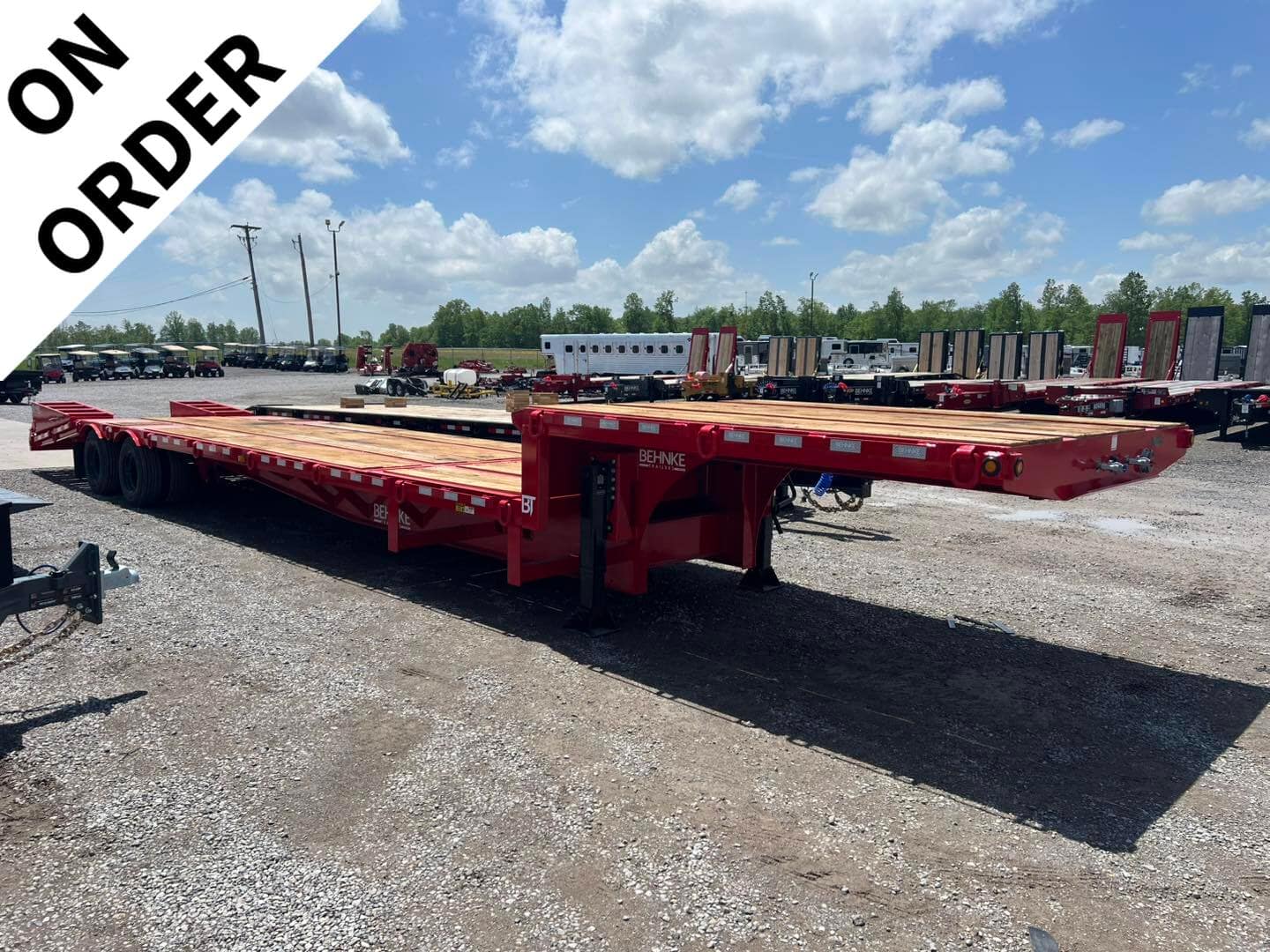 Red Behnke flatbed trailer with a long wooden deck in an outdoor equipment yard, a white diagonal 'ON ORDER' banner across the corner.