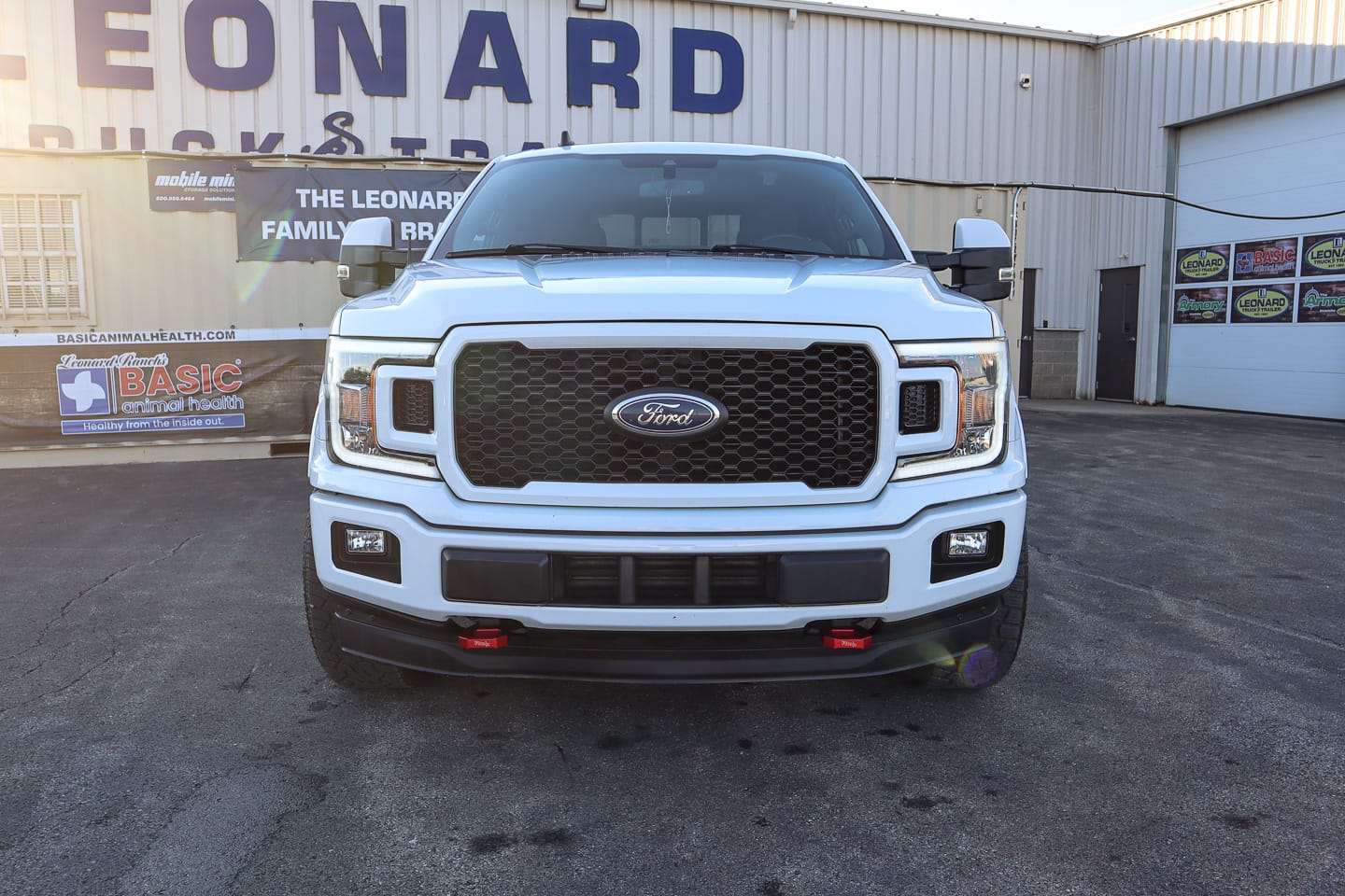 Front view of a white Ford pickup truck parked in a dealership lot with a large LEONARD sign in the background.