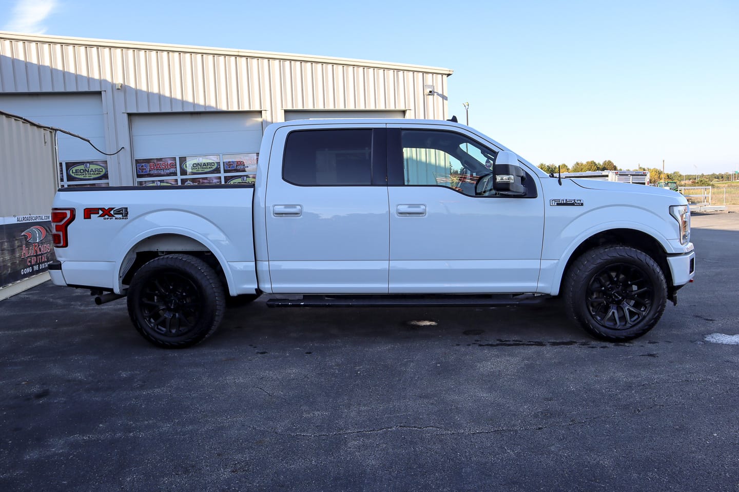 Side view of a white Ford F-150 pickup with black wheels parked in front of a metal industrial building