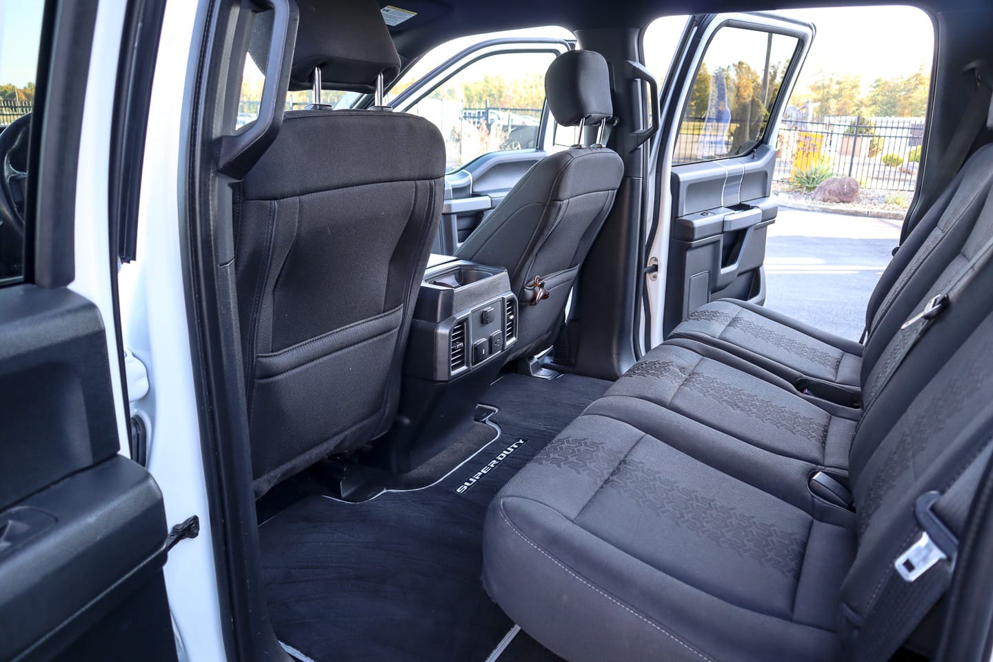 Interior of a white pickup truck, showing the back bench seat, front seats, and center console with the rear door open to a parking lot outside.