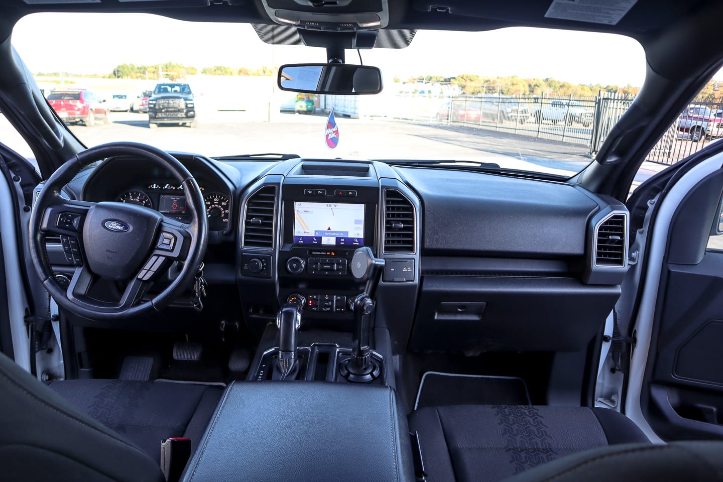 Interior view of a dark pickup cabin from the back seat: steering wheel on the left, large central touchscreen, gear shifter, and black cloth seats.
