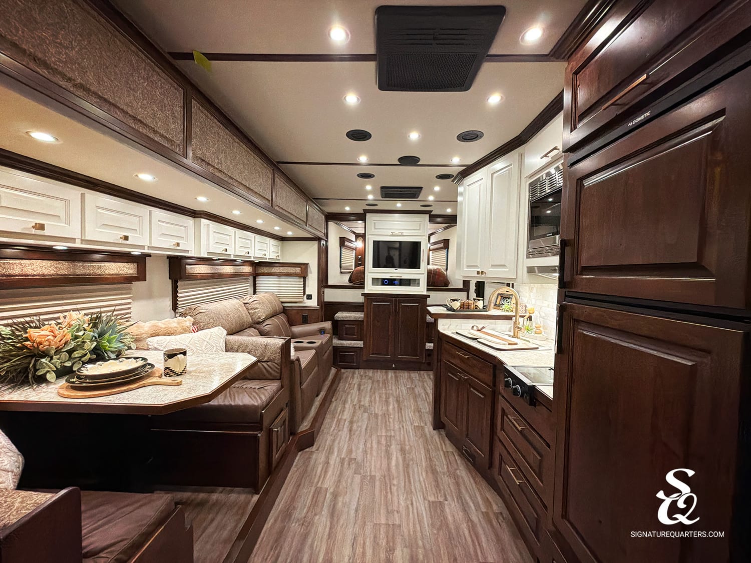 Luxurious RV interior with dark wood cabinets, beige sofa, and a central entertainment nook under a mounted TV and ceiling lights.