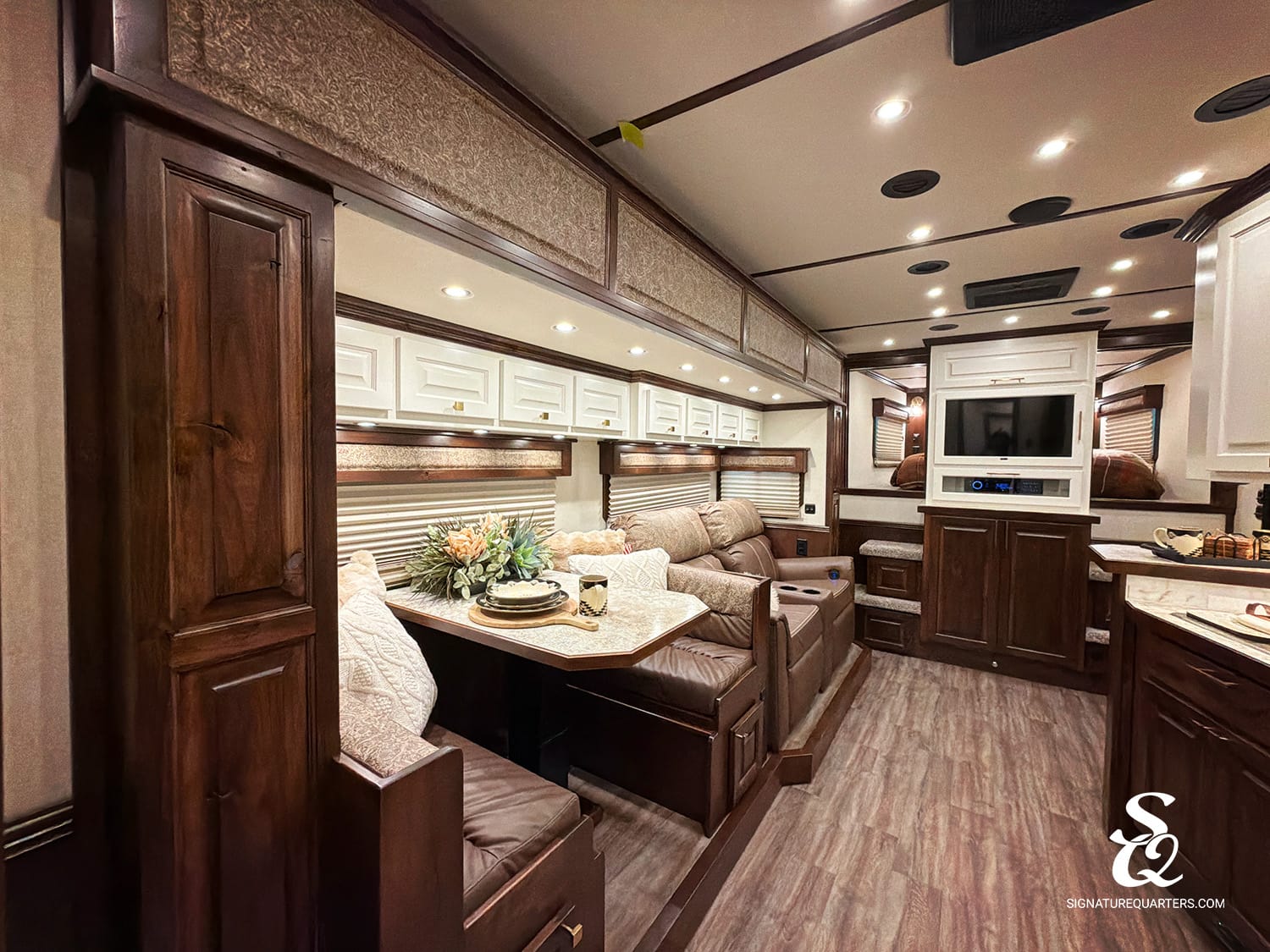 Luxurious motorhome interior with dark wood cabinets, beige upholstery, and a booth dinette beside a windowed wall setup.