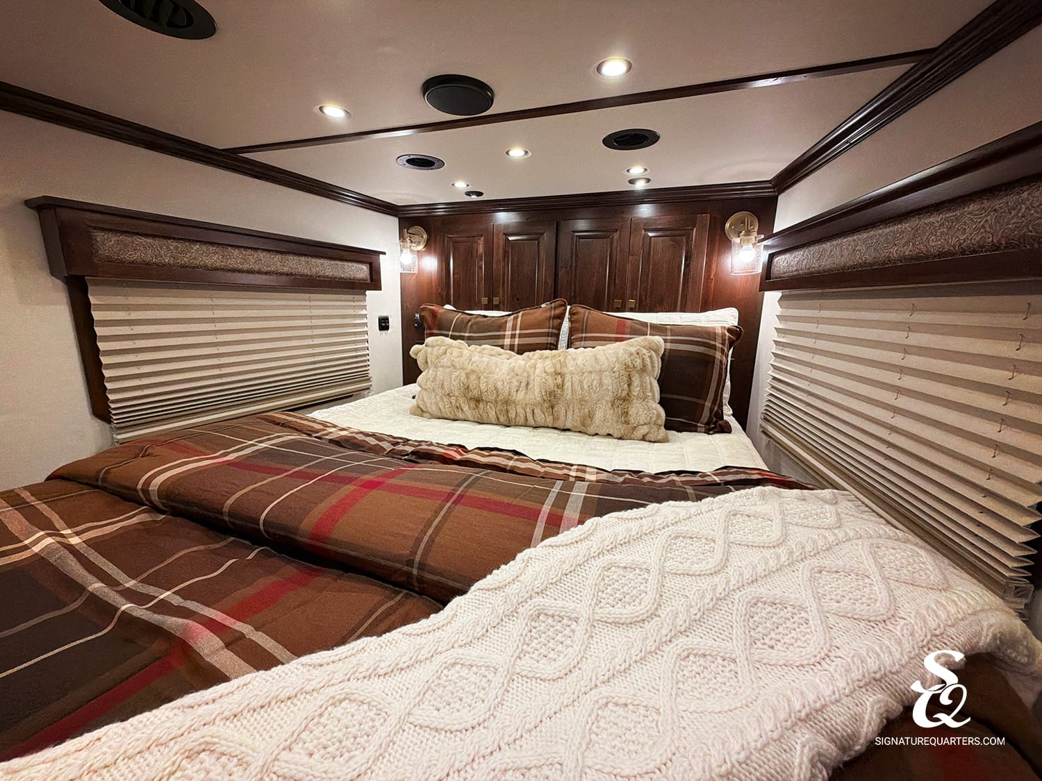 Cozy RV bedroom with a large bed, plaid brown bedding, and a cream knitted throw against dark wood cabinetry on the head wall.