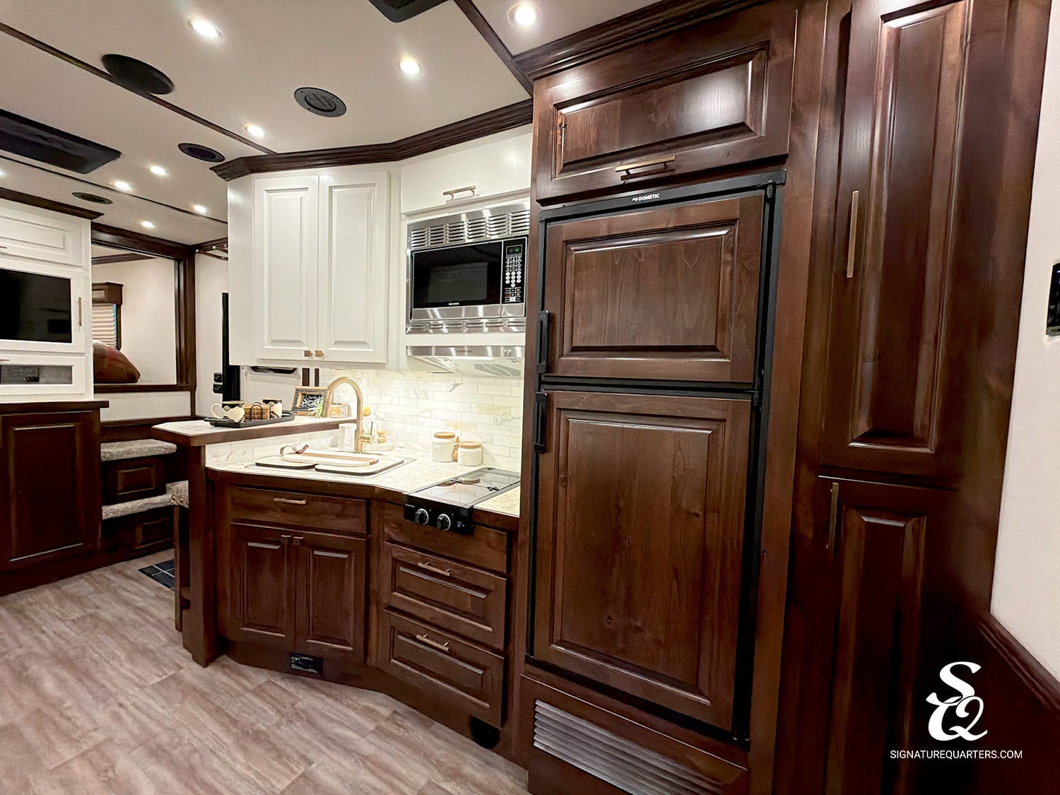 Luxurious RV kitchen with dark wood cabinets, white upper cabinets, stainless microwave, and a double refrigerator surrounding a beige countertop and sink area.