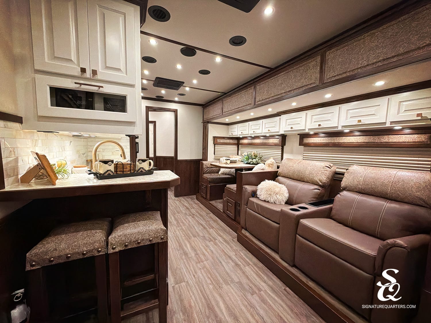 Luxurious RV interior with a kitchenette on the left, brown leather recliners on the right, white cabinets, and warm recessed lighting.