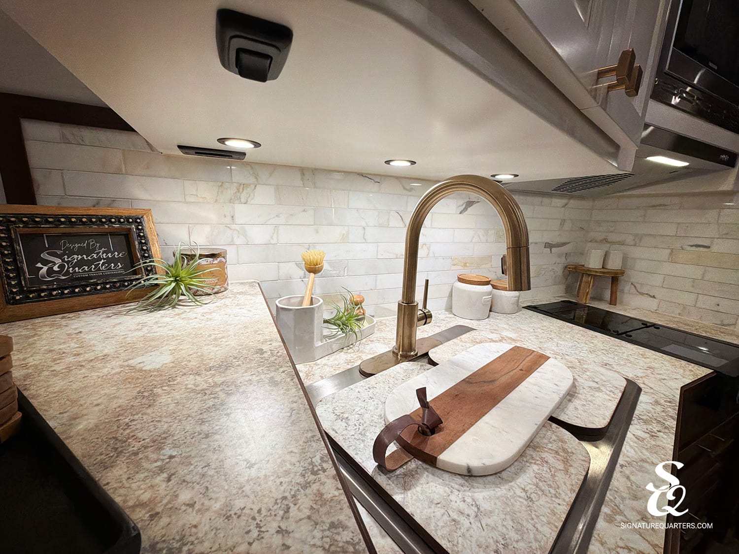 Modern kitchen counter scene with a curved brass faucet, cutting board, and small potted plants on a marble countertop plate.”
