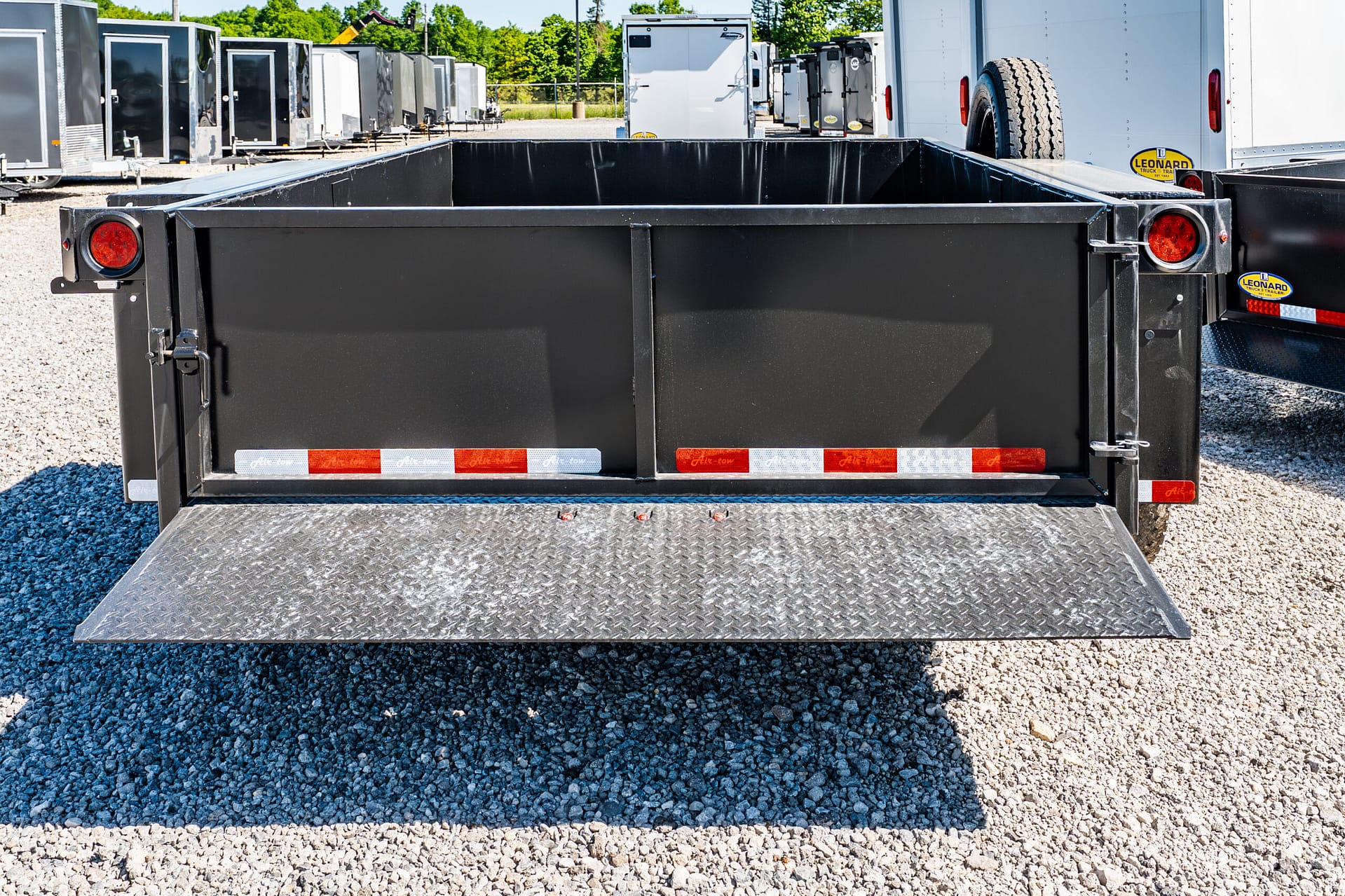 Black open-top trailer with a lowered diamond-plate ramp, parked among other trailers in a gravel lot.