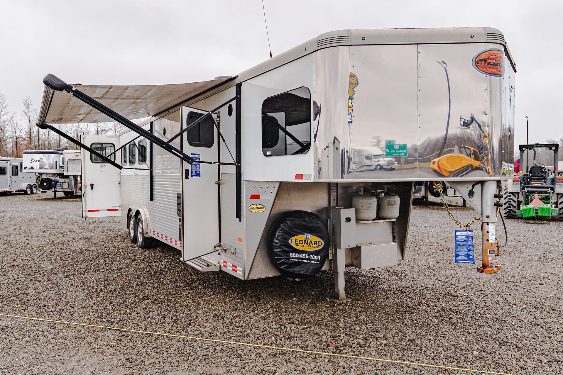 Large silver RV trailer with a deployed awning and open side entry on a gravel lot; other trailers visible in the background.