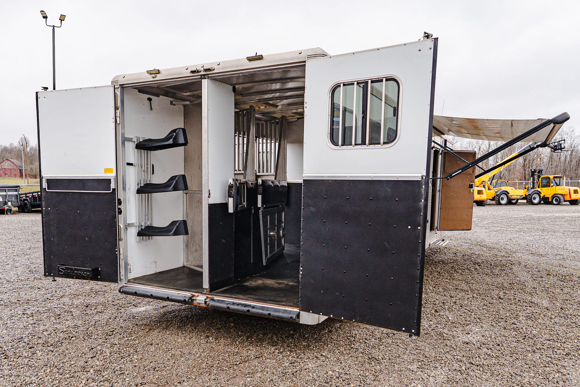 Open horse trailer with divider stalls and seating visible inside, doors swung wide on a gravel lot; construction equipment in the background.