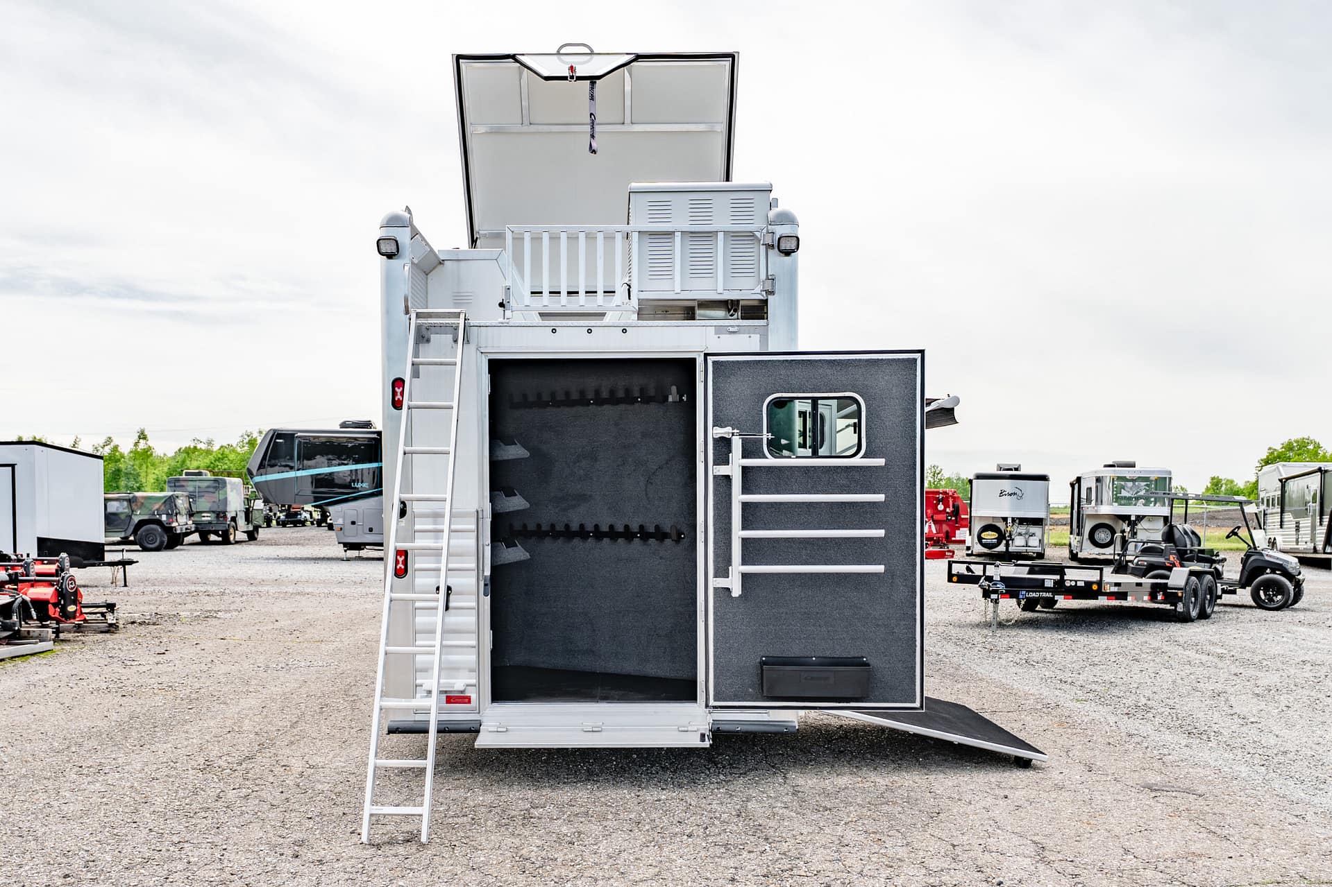 Rear view of a large white trailer with an open hatch, ladder down, and a black interior, parked in a gravel lot with other trailers nearby.