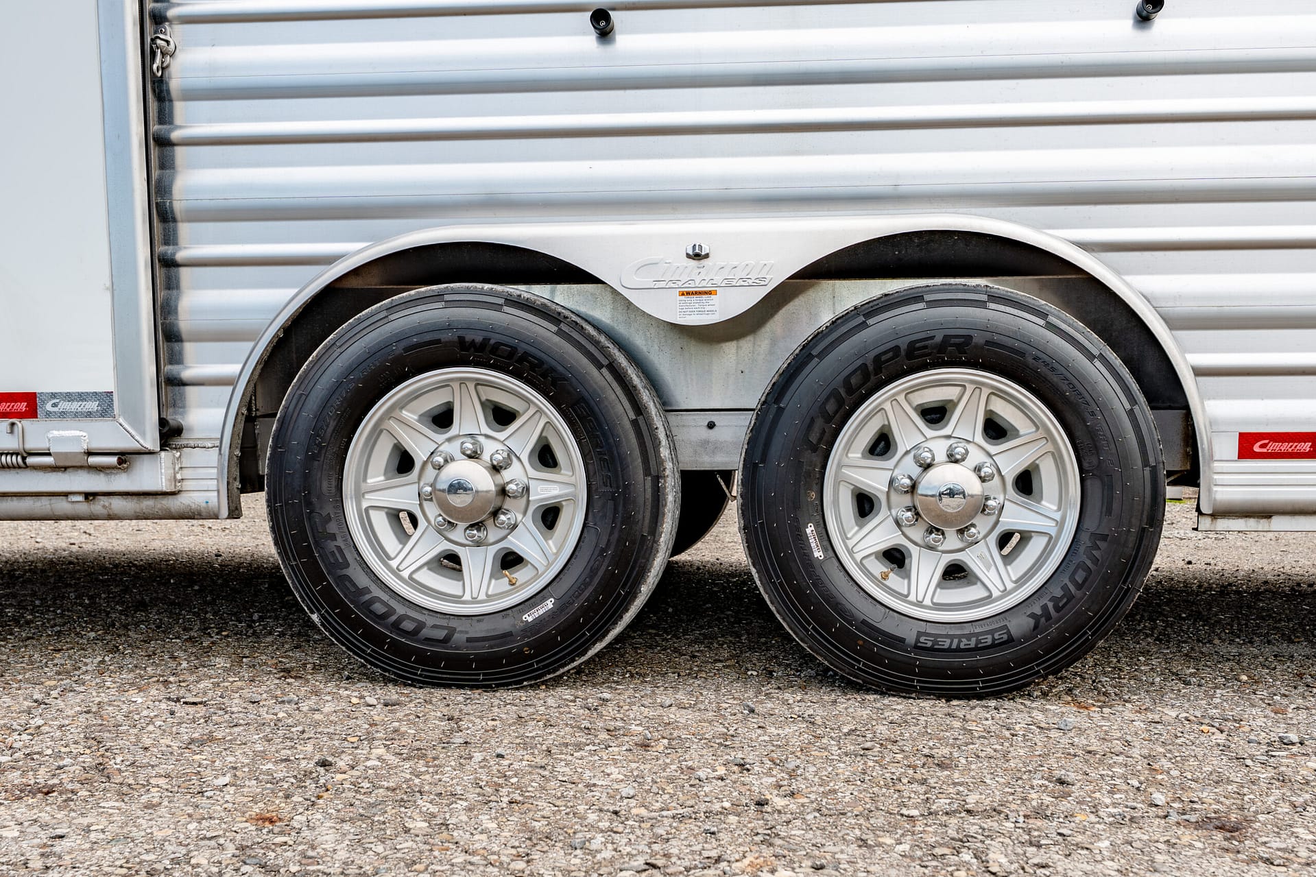 Side view of a silver trailer with dual wheels and chrome rims, on a gravel surface.