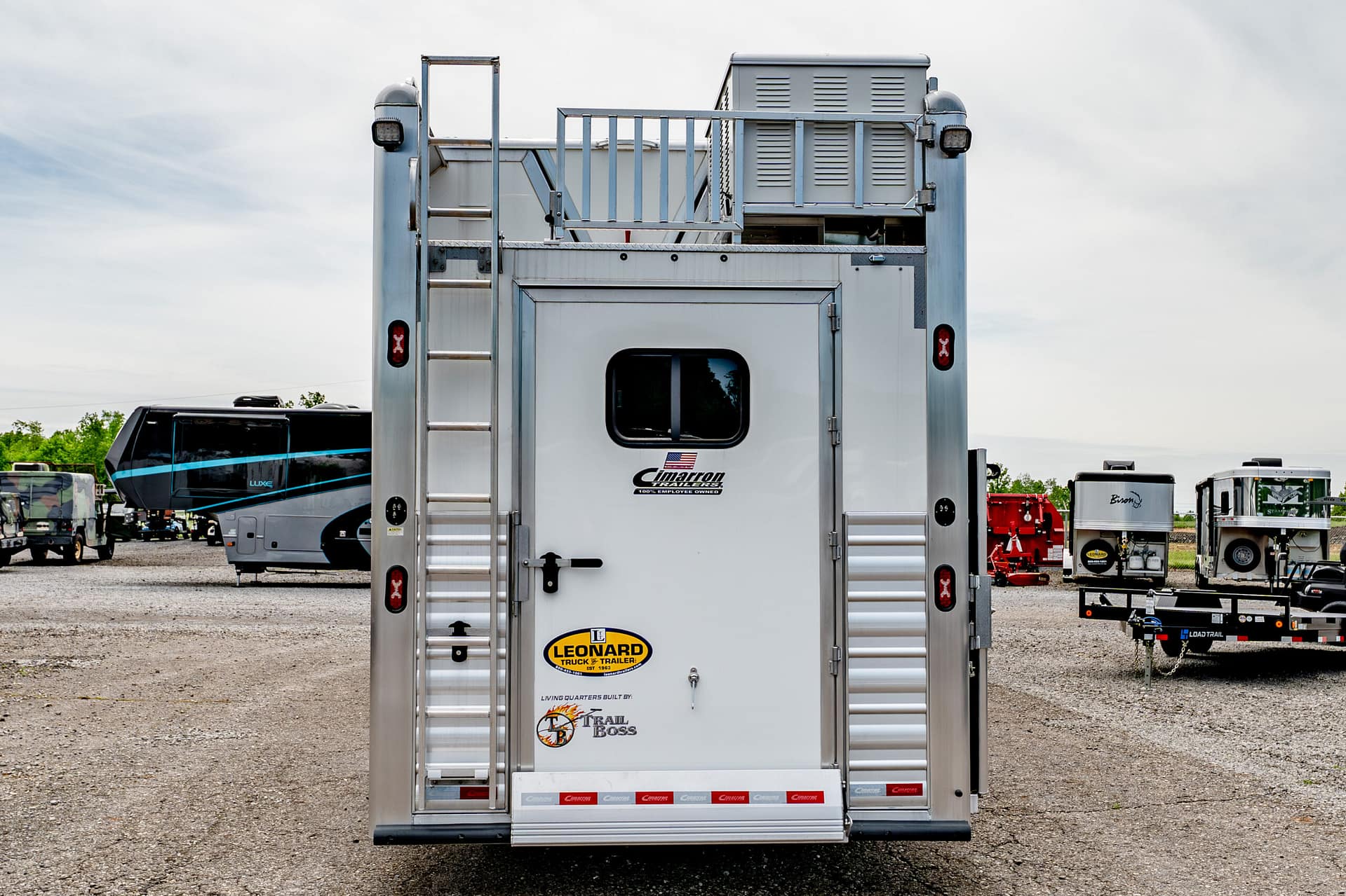 Rear view of a silver trailer with a ladder on the left, vents, and branding decals, parked in a trailer yard with other vehicles nearby.