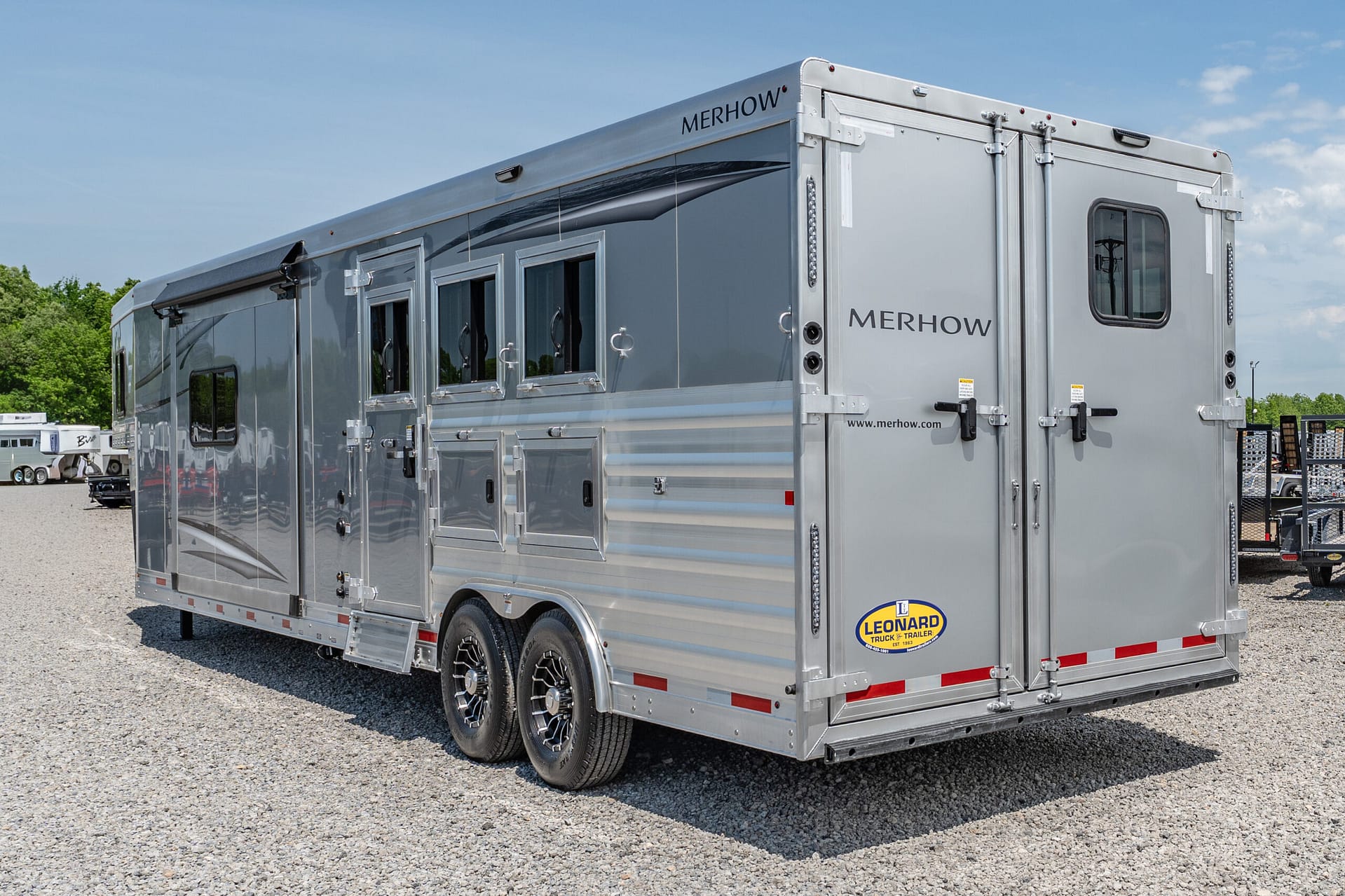 Large silver Merhow horse trailer with side windows, parked on a gravel lot under a blue sky.