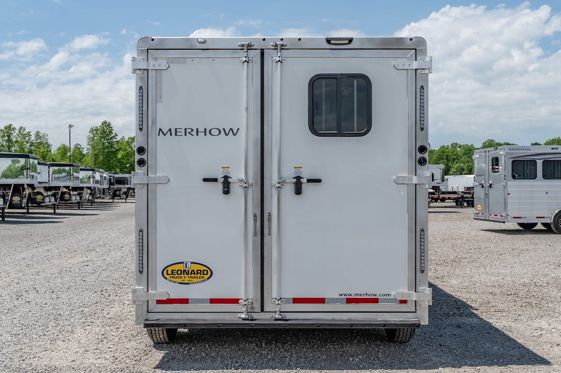 Rear view of a silver Merhow trailer with dual doors and a small window, parked on a gravel lot with other trailers in the background.