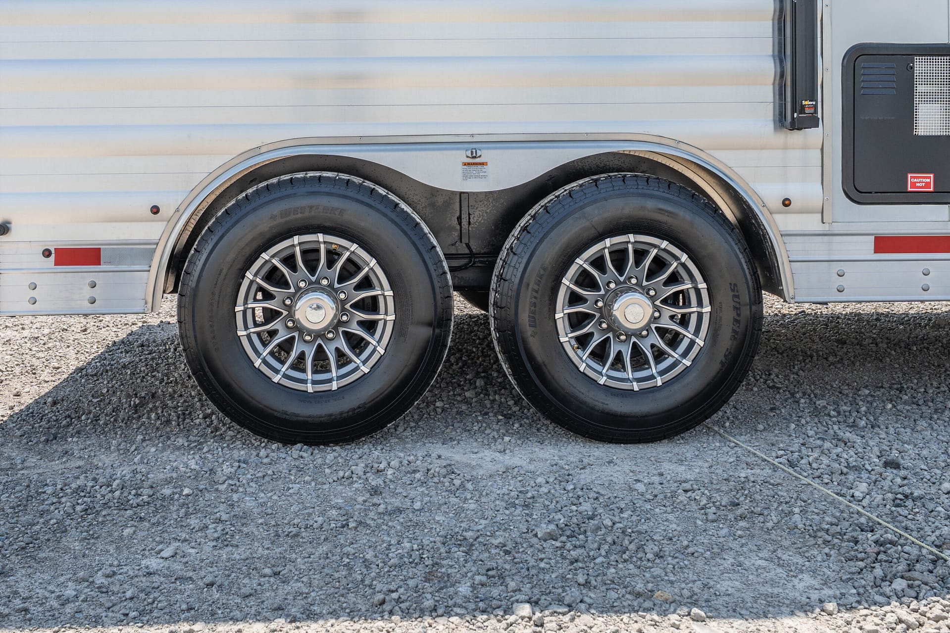 Side view of a metal trailer with twin wheels and a curved wheel well over a gravel surface, showing chrome rims and tire treads
