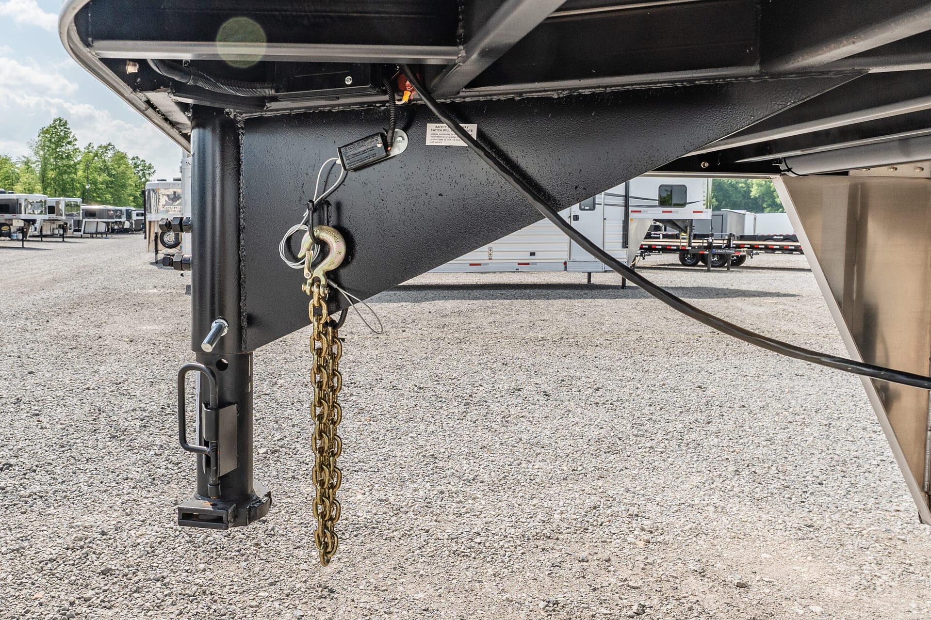 Close-up of a trailer hitch with safety chains and hitch pin, undercarriage in black metal, on a gravel lot with other trailers in the background.