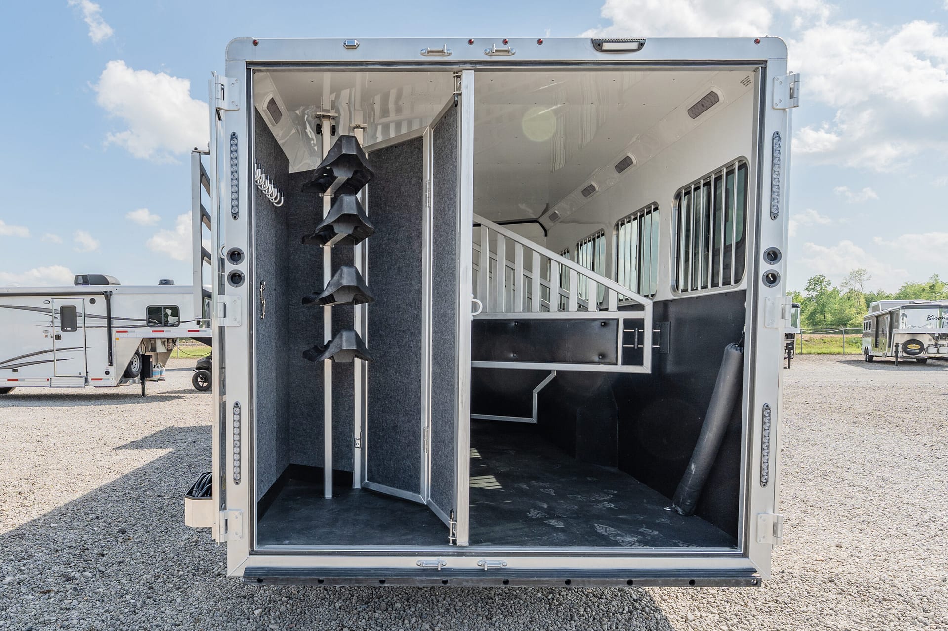 Open rear of a silver horse trailer showing interior stalls, tack racks on the left, and windows along the right side.